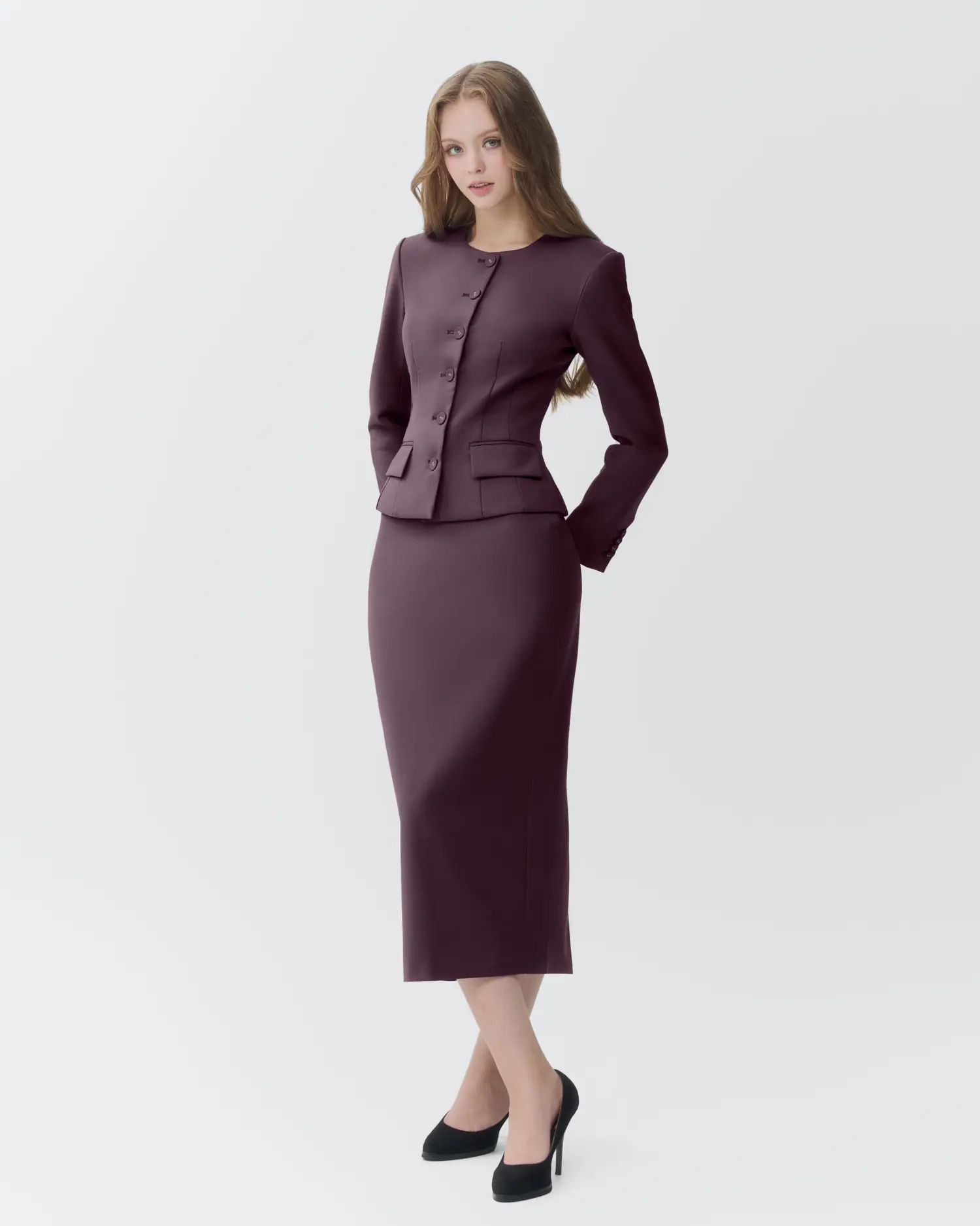 Woman in burgundy suit and black heels posing against a white background.