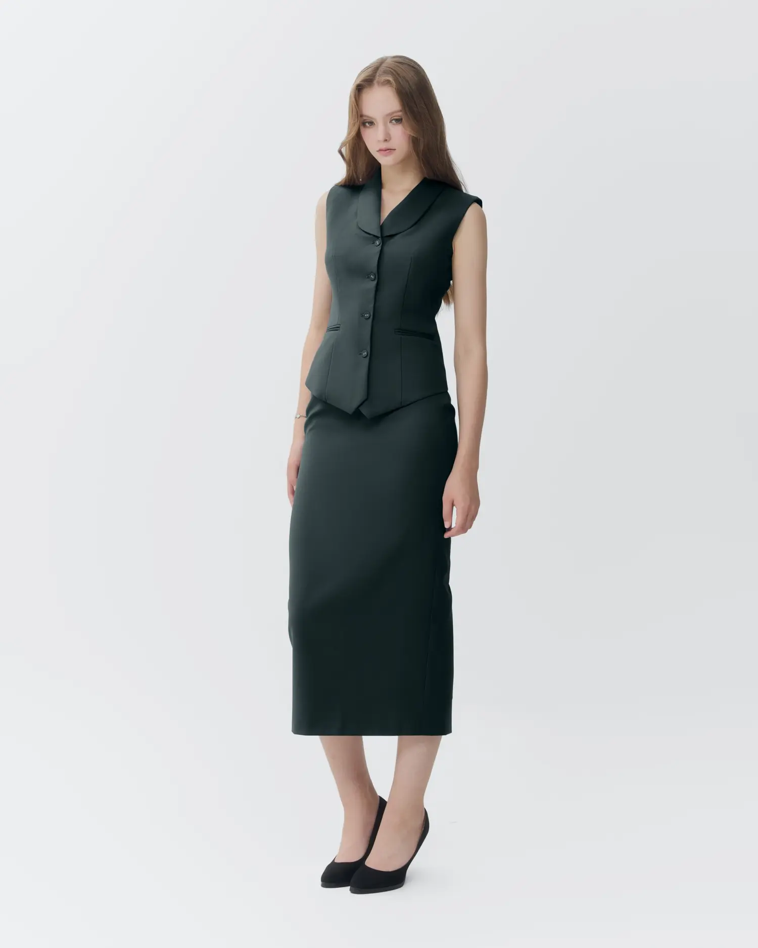 Woman in stylish dark green vest and skirt outfit against a plain background, showcasing modern fashion elegance.