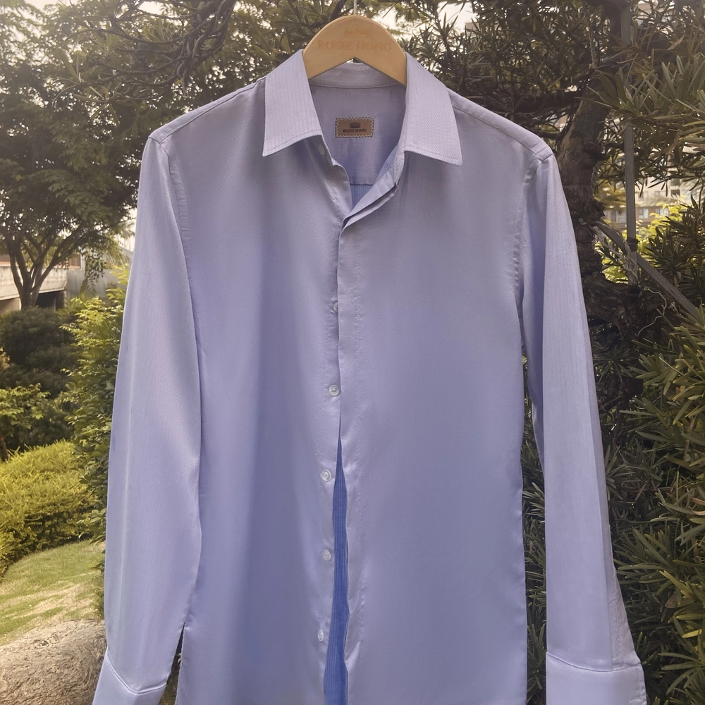 Light blue dress shirt on hanger outdoors with green foliage background.