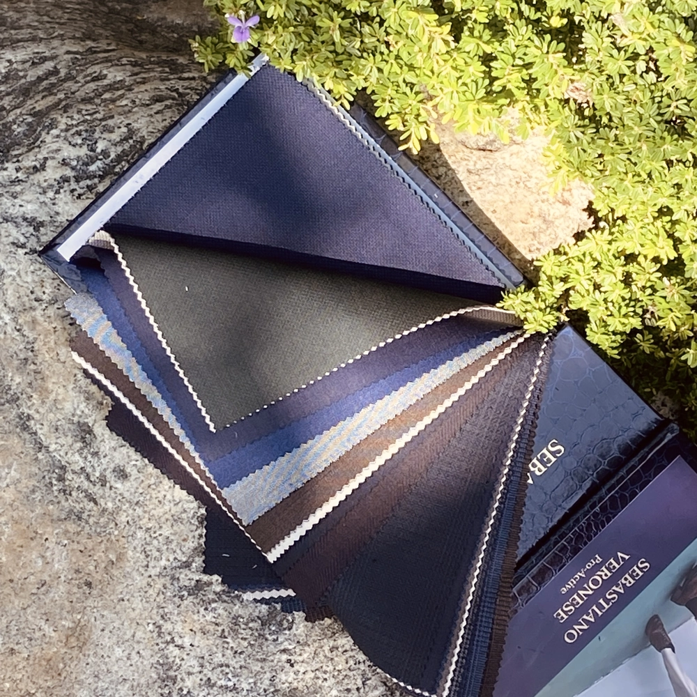 Fabric swatches in various shades of blue and gray on a stone surface, surrounded by greenery.