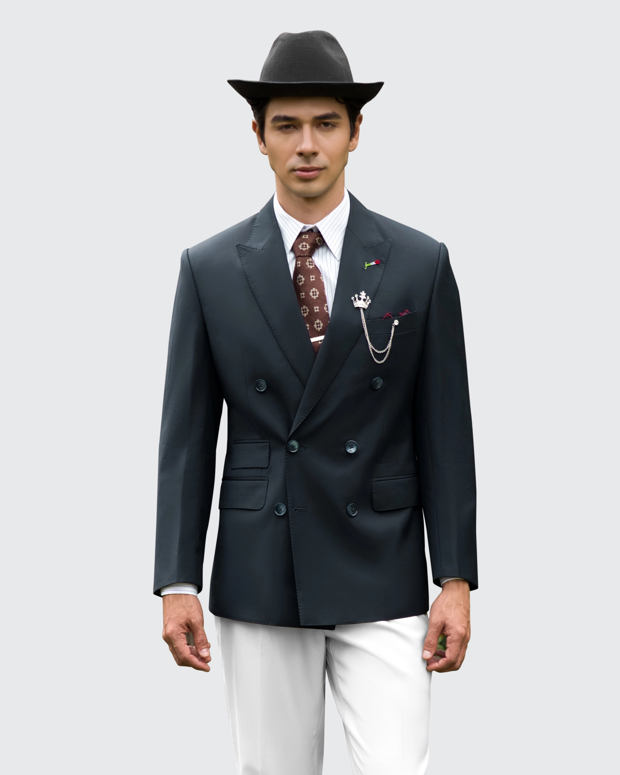 Man in a stylish black suit and hat with a patterned tie and lapel pin, standing confidently against a gray background.