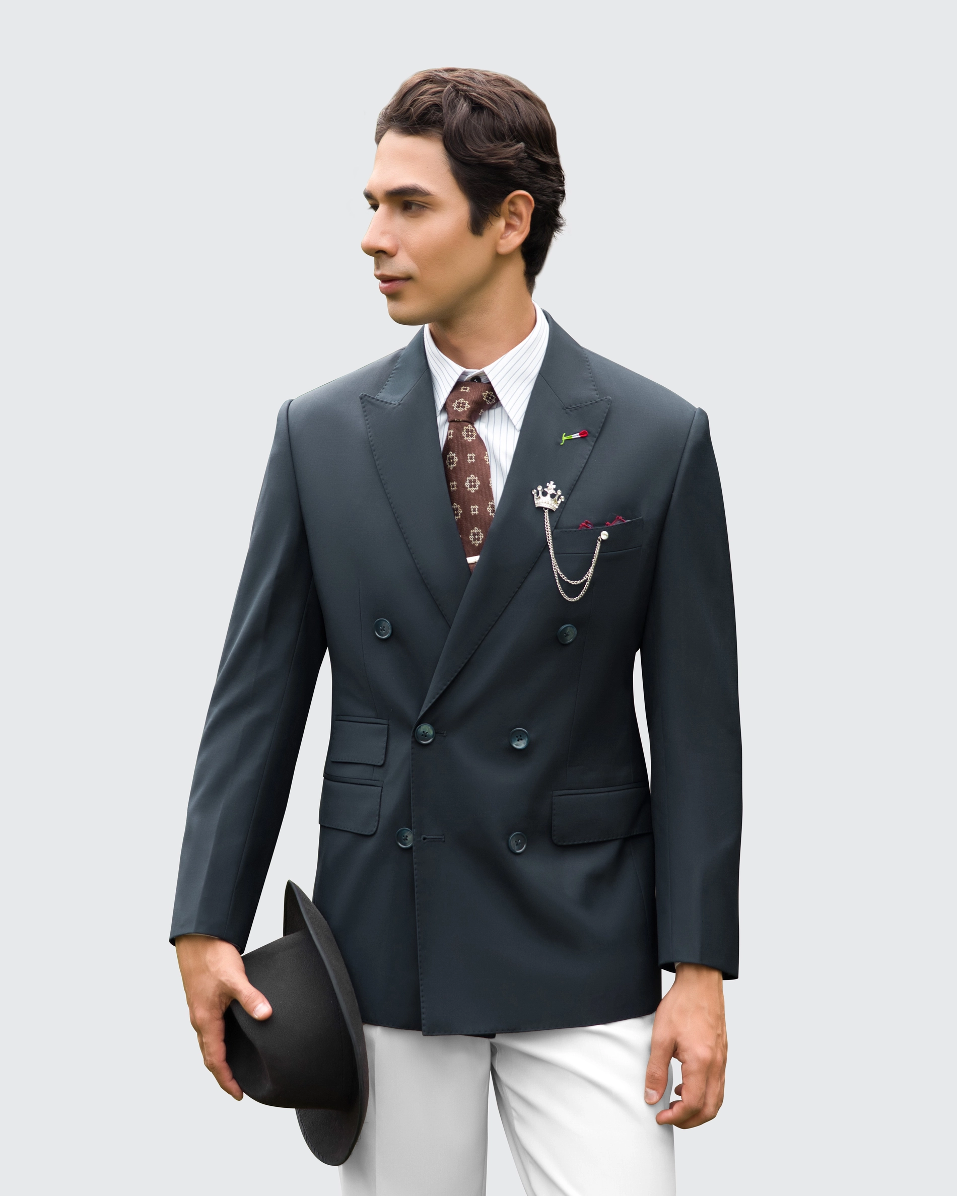 Man in tailored suit with accessories and holding a hat, looking left, stylish and elegant formal attire.