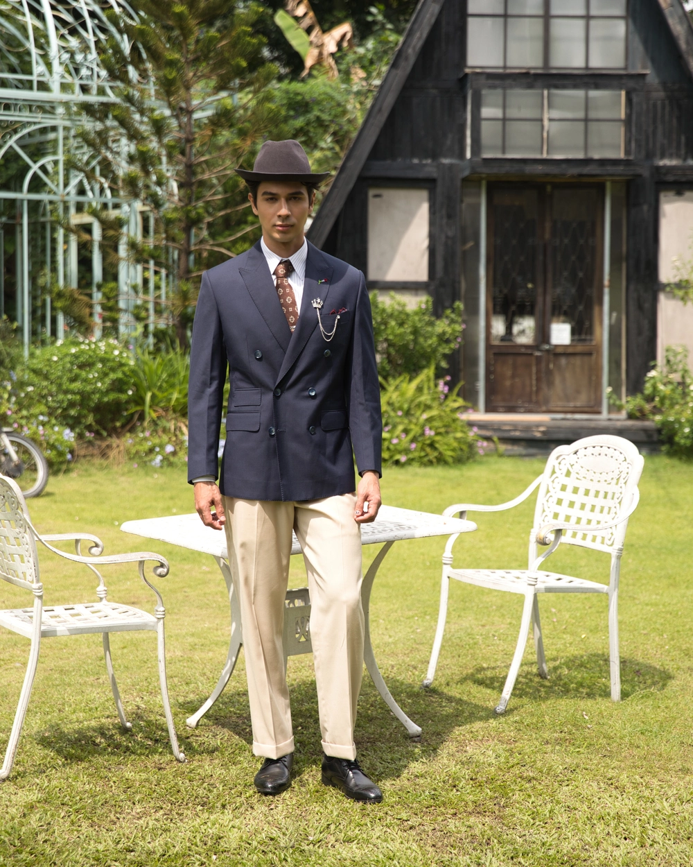 Man in vintage suit and hat stands near garden table and chairs outside a quaint house.