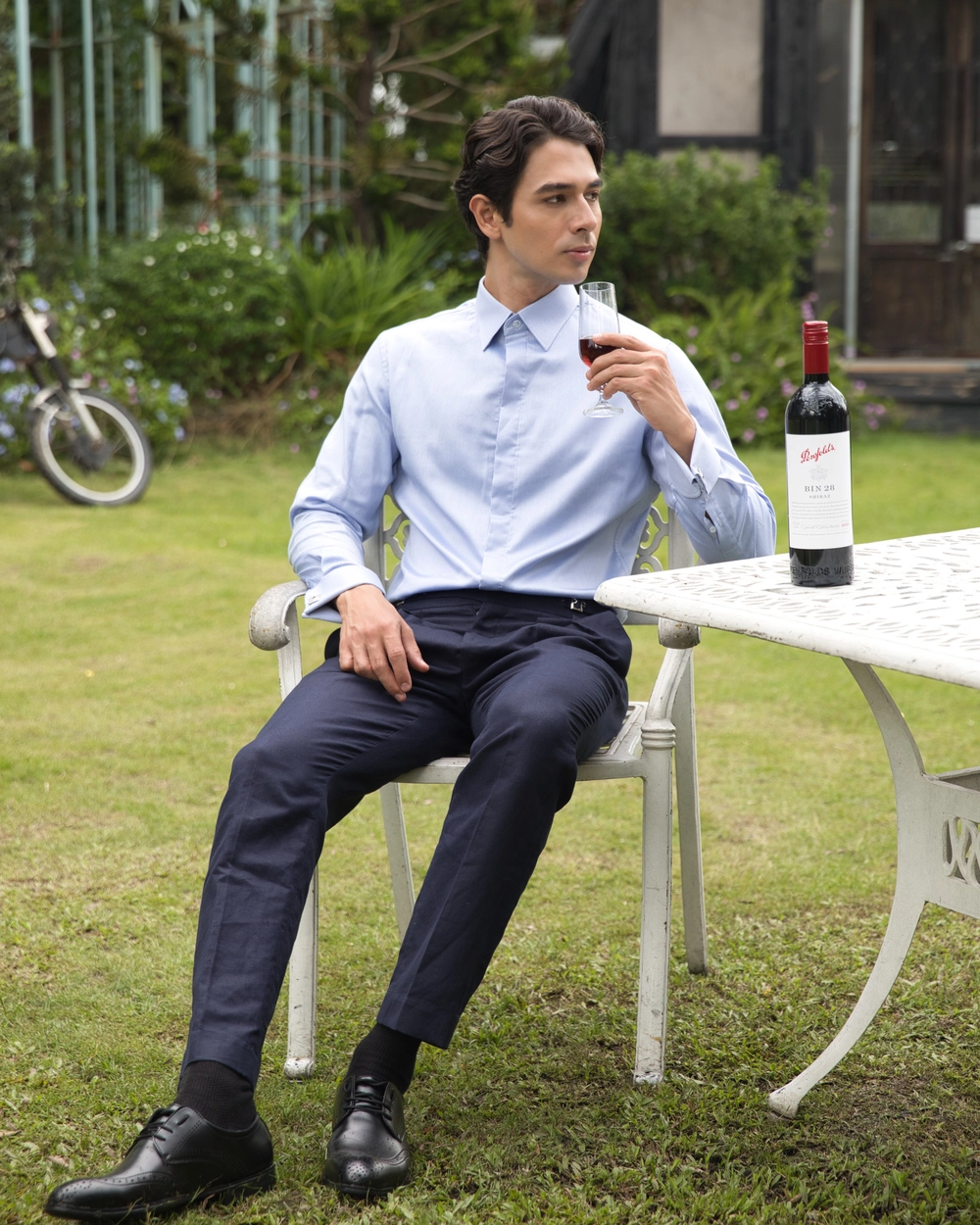 Man in a blue shirt enjoying wine outdoors, sitting by a patio table in a lush garden setting.
