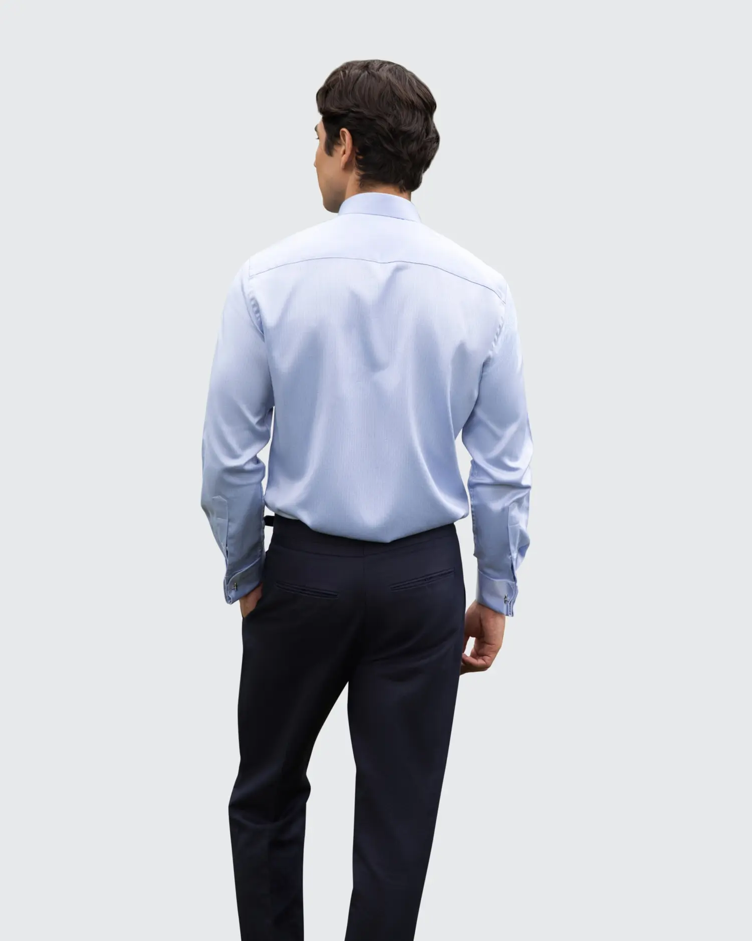 Man in light blue dress shirt and dark pants, standing with hands in pockets, back view on a gray background.