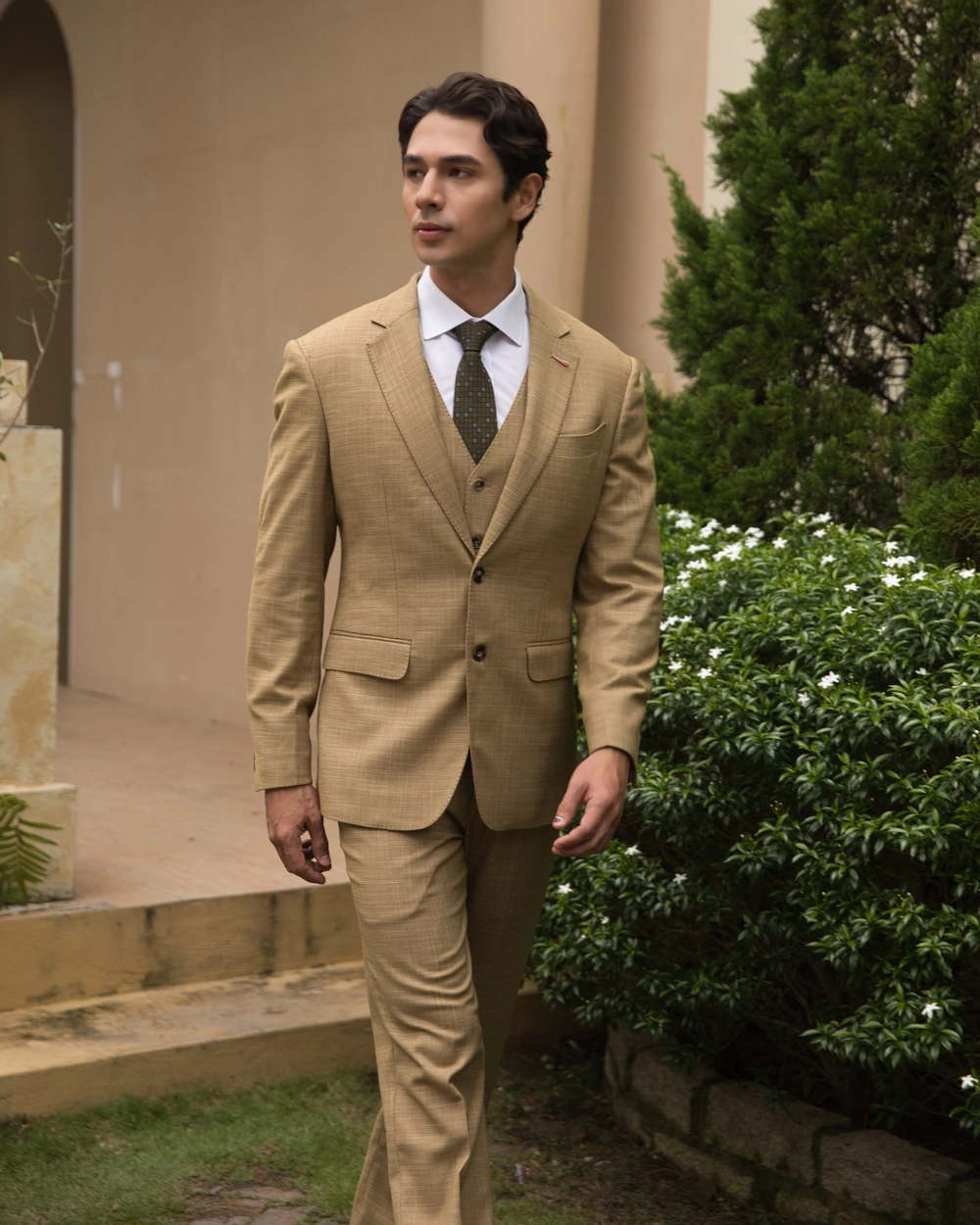 Man in a beige suit walking outdoors, surrounded by greenery and a modern building facade.