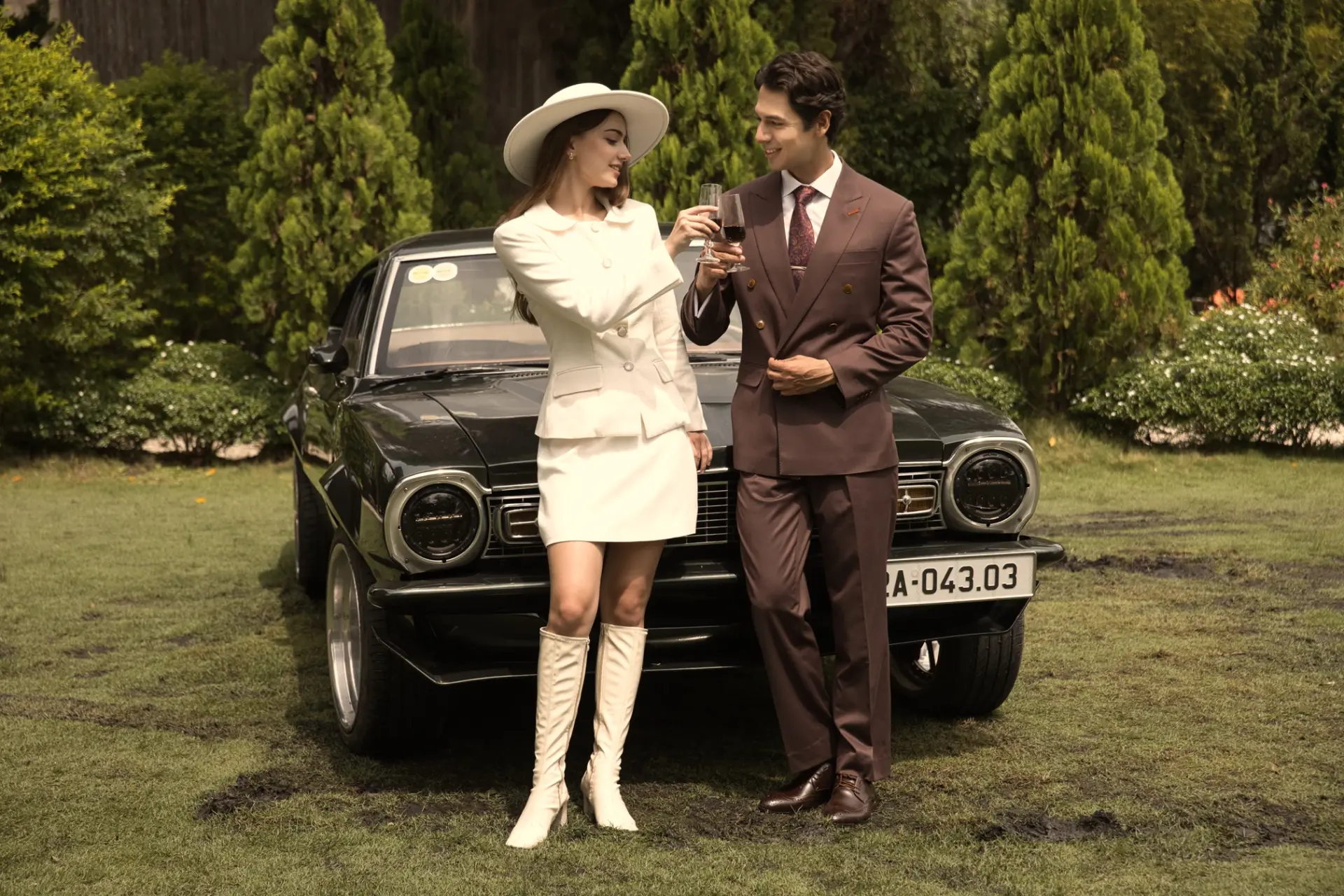 Couple toasting with wine beside a vintage car in a garden setting, dressed in elegant attire.