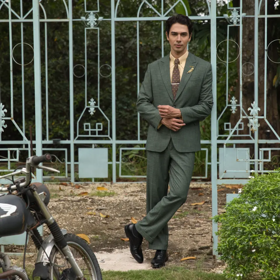 Man in green suit standing by vintage gate, next to an old motorcycle, outdoors.
