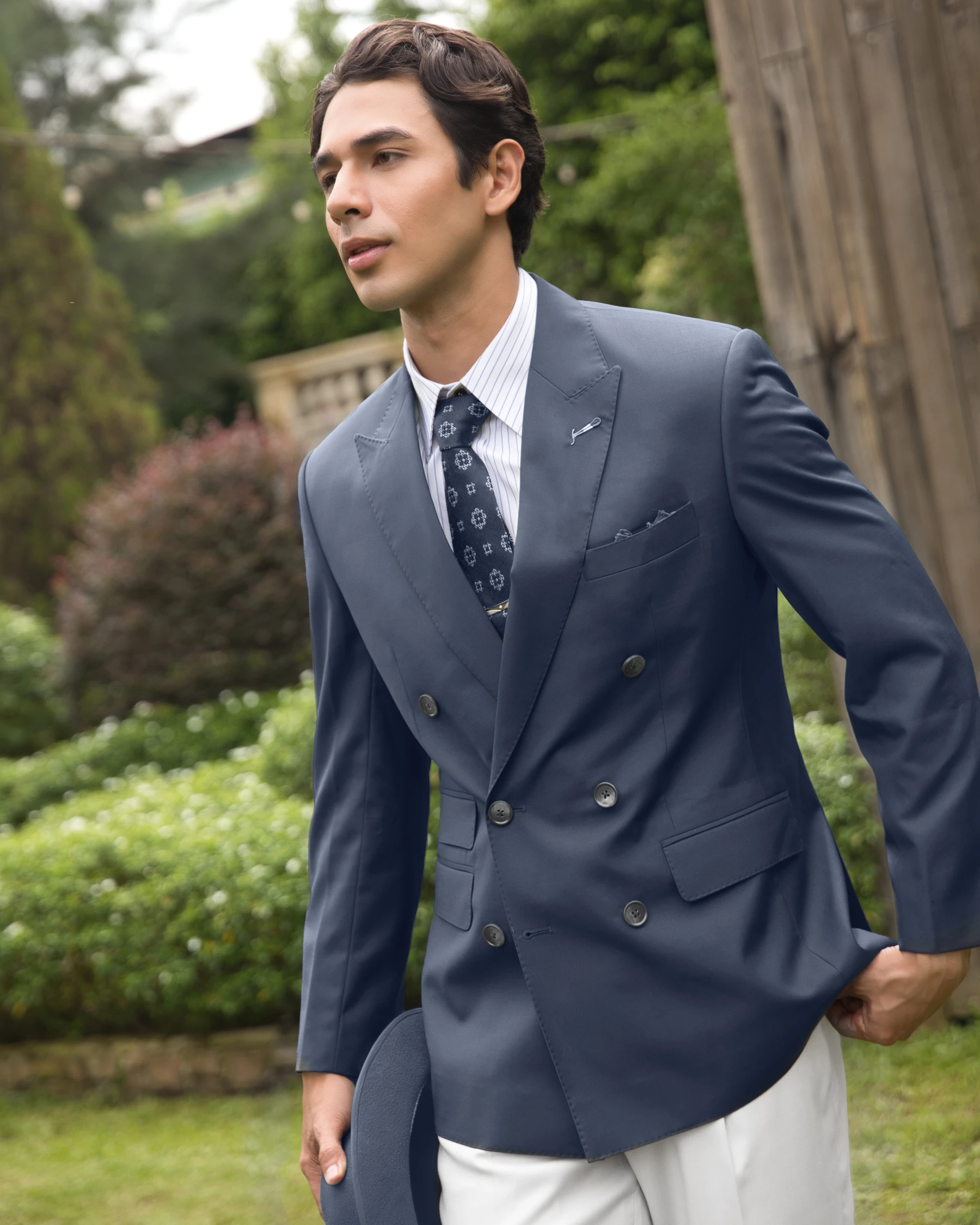 Man in stylish blue suit with tie walking in a garden, holding a blue hat, surrounded by greenery.