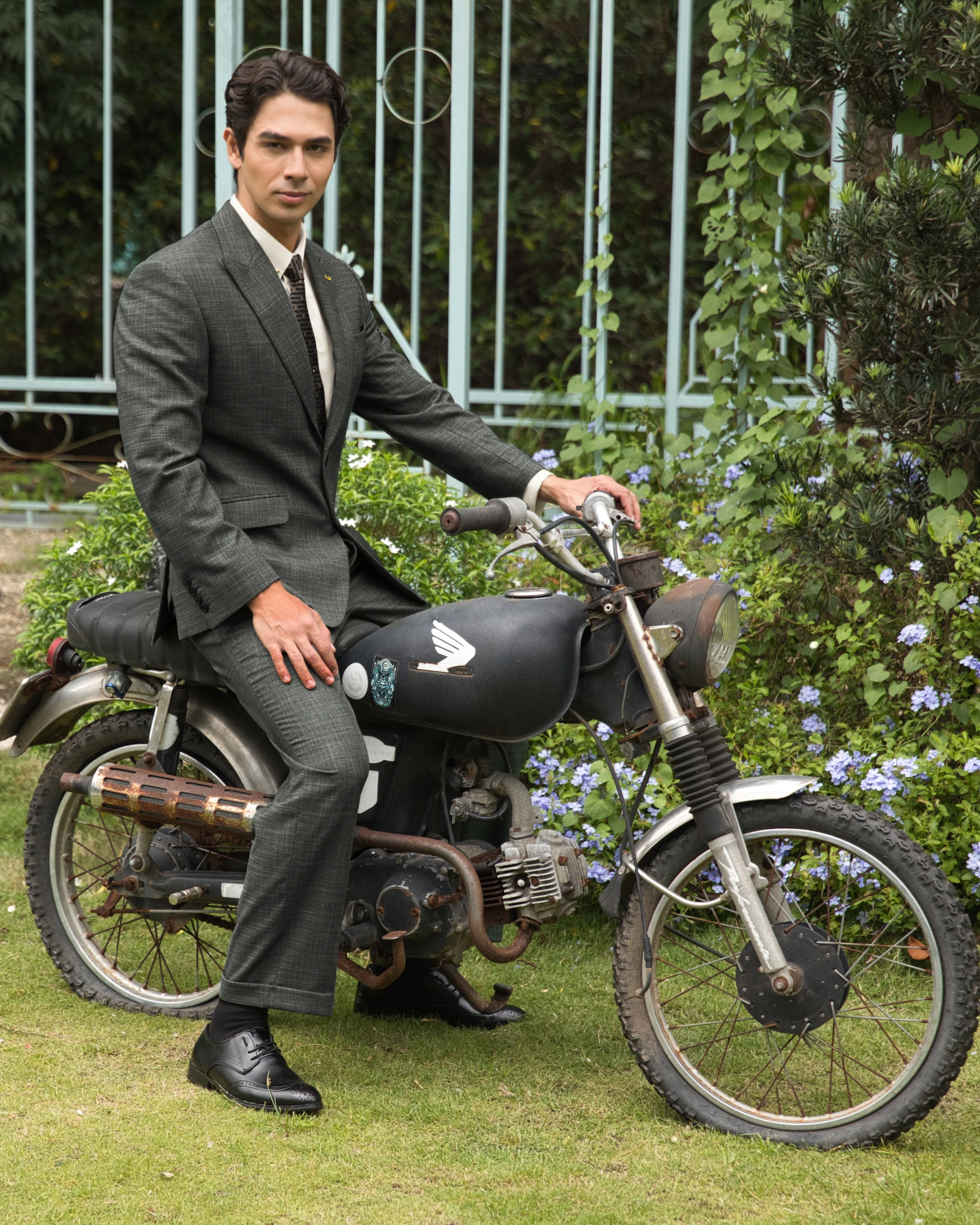 Man in a suit sitting on a vintage motorcycle in a garden setting with green foliage and flowers.