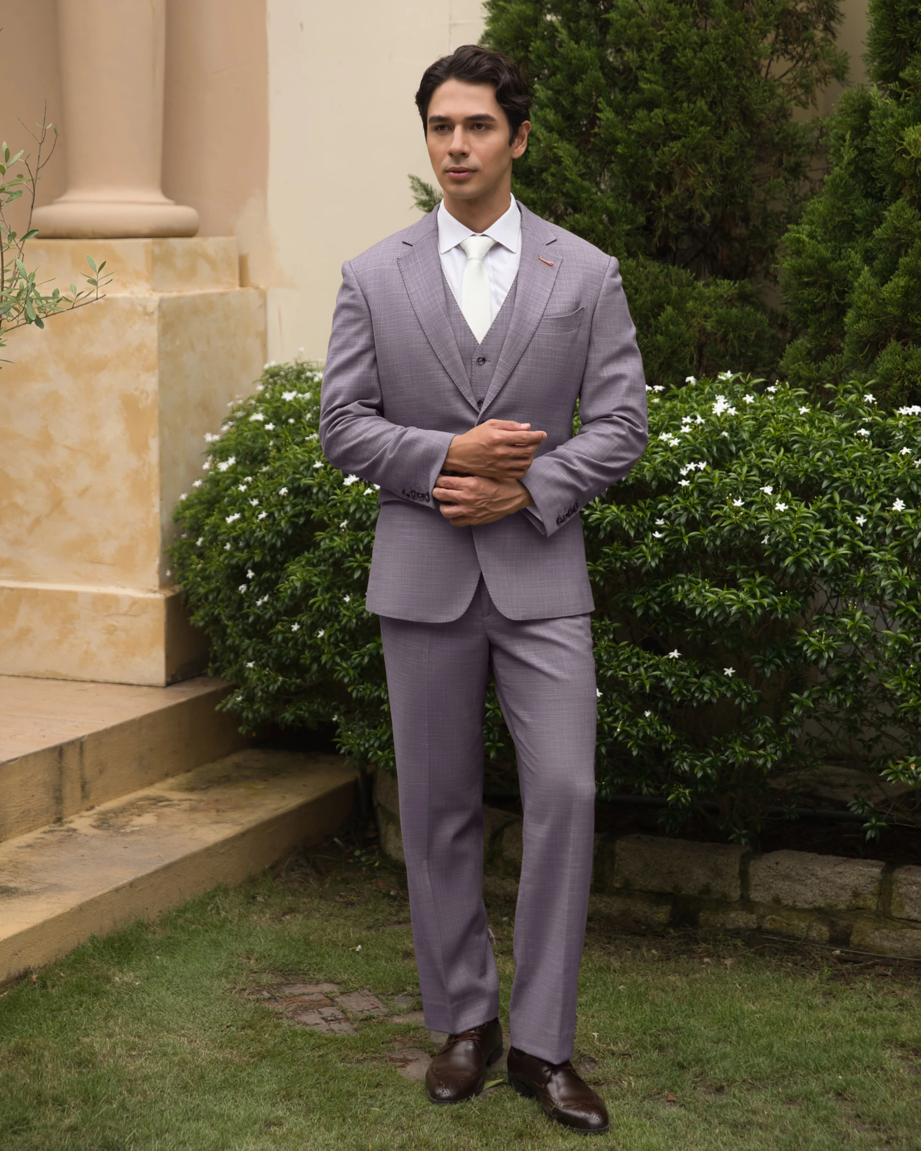 Man in a gray suit standing on grass near green bushes and stone steps, looking confident.