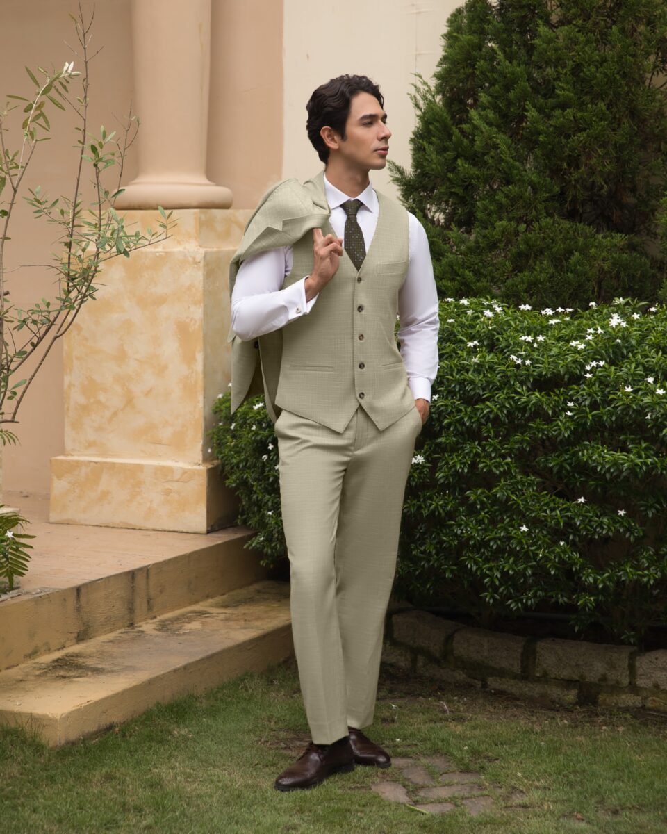 Man in light suit leans against a pillar, holding jacket over shoulder, surrounded by greenery.
