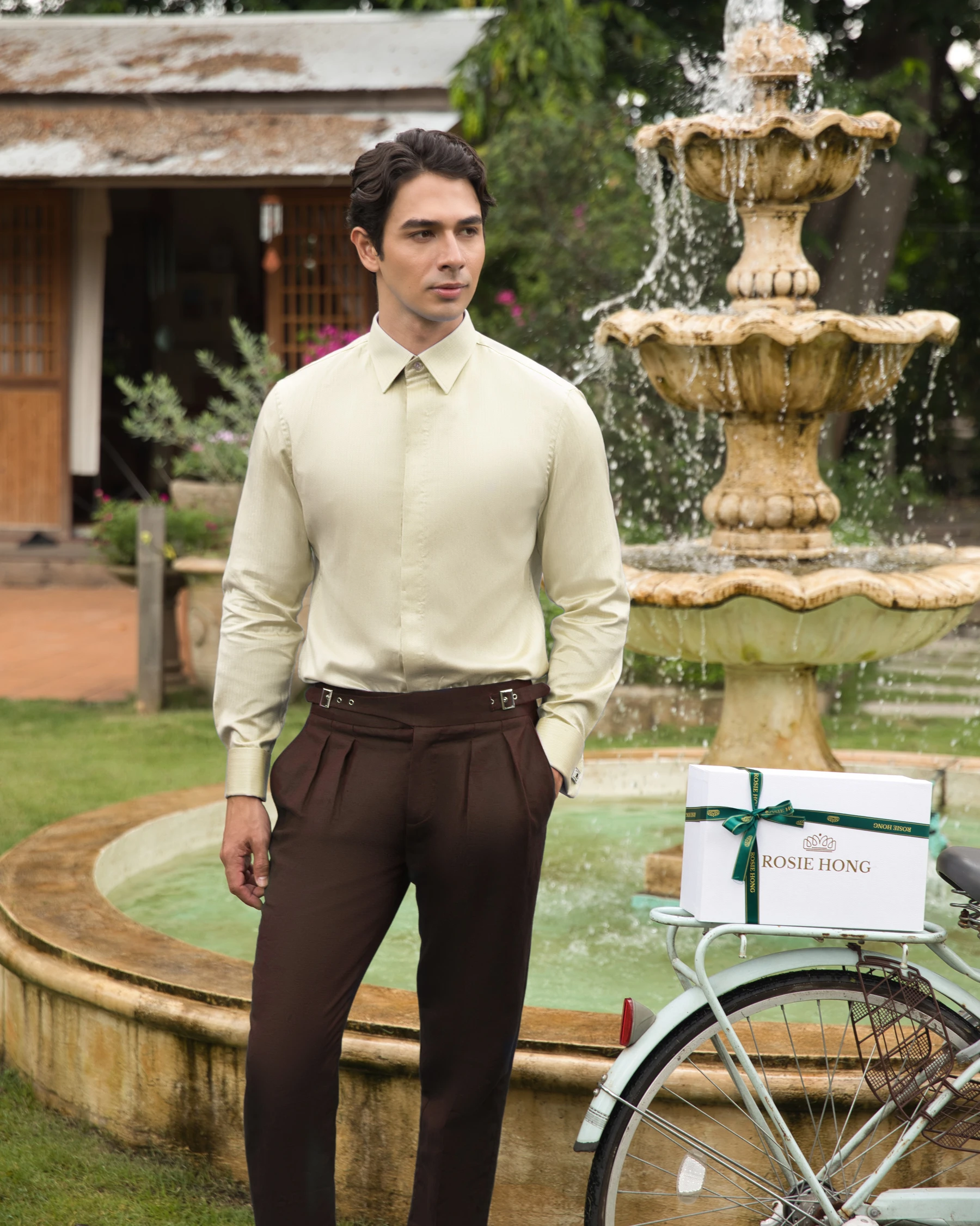 Man in formal attire stands by a fountain, beside a bicycle with a gift box labeled Rosie Hong.