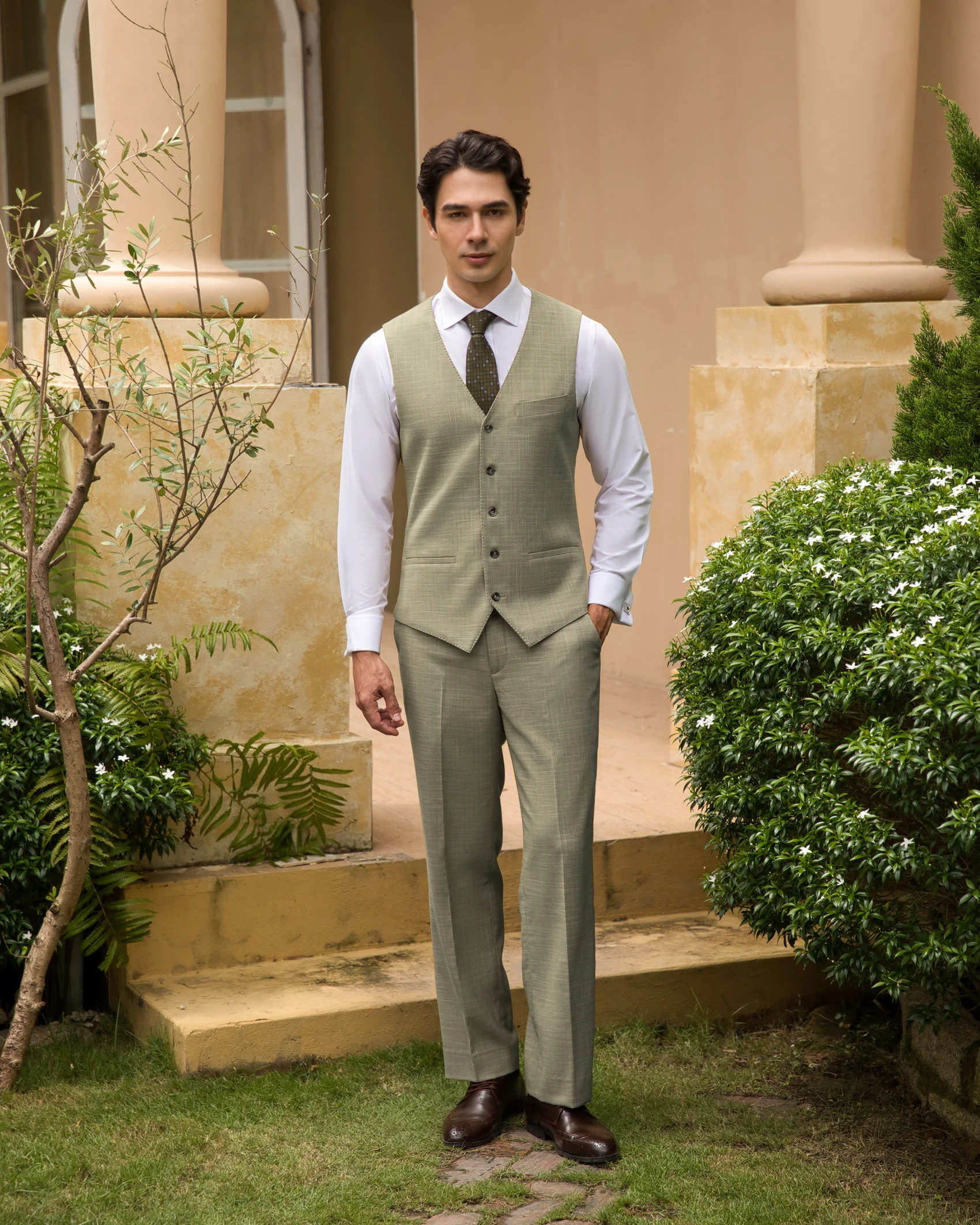 Man in a light green suit standing outdoors next to greenery and a building, looking directly at the camera.