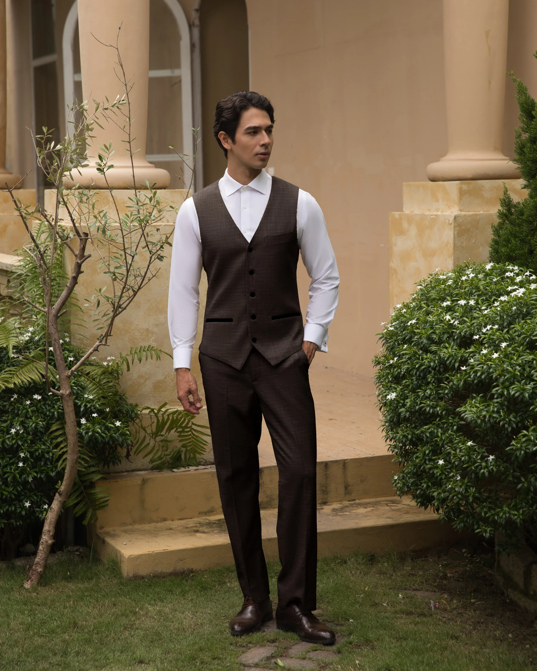 Man in a brown suit vest and white shirt stands outside by garden and columns, exuding a sophisticated style.