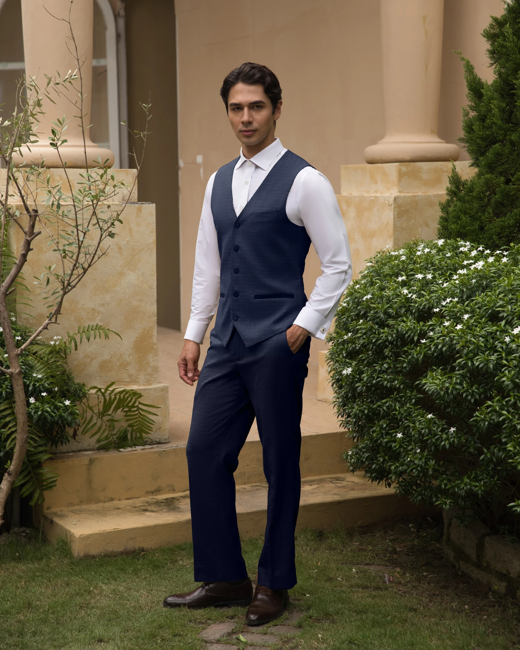 Man in navy vest and trousers standing outside a building, surrounded by greenery.