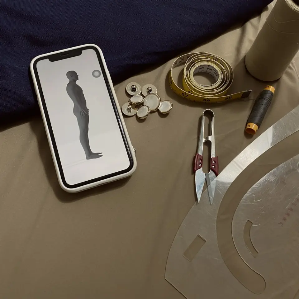 Sewing tools laid out beside a smartphone showing a 3D body scan model, illustrating the blend of traditional tailoring and modern scanning technology.