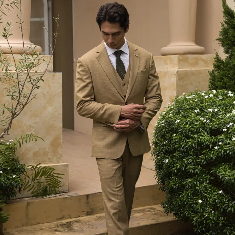 Man in brown suit and tie standing by ornate columns and lush green bushes, exuding elegance and sophistication.