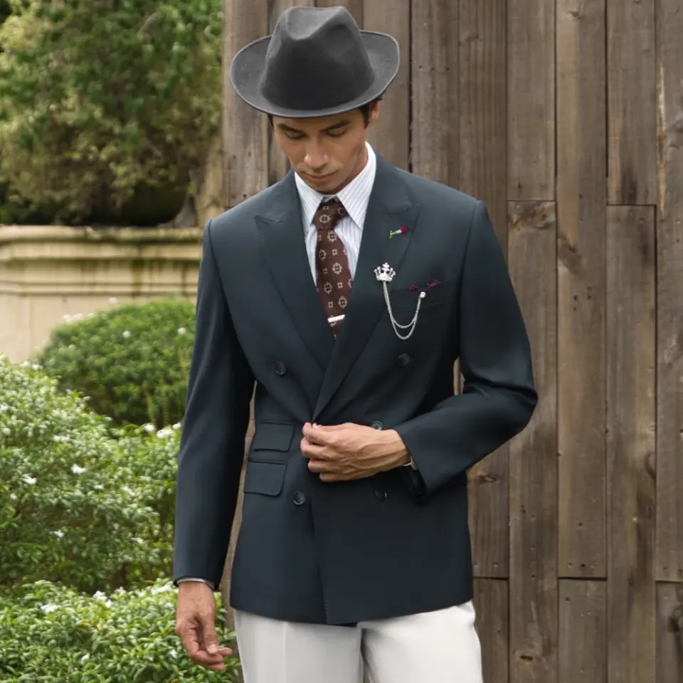 Man in vintage suit and hat stands in front of wooden wall and greenery, exuding classic style and elegance.