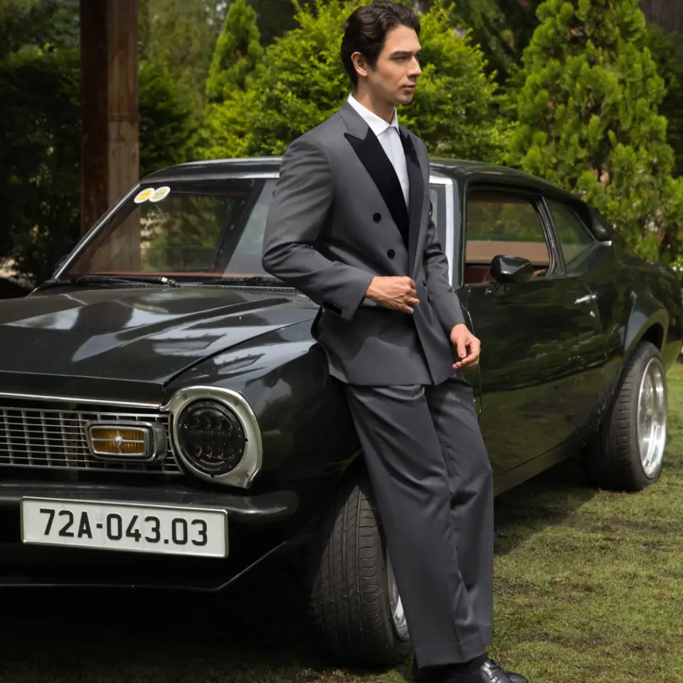 Man in a suit leaning on a vintage car, set in a lush garden backdrop.