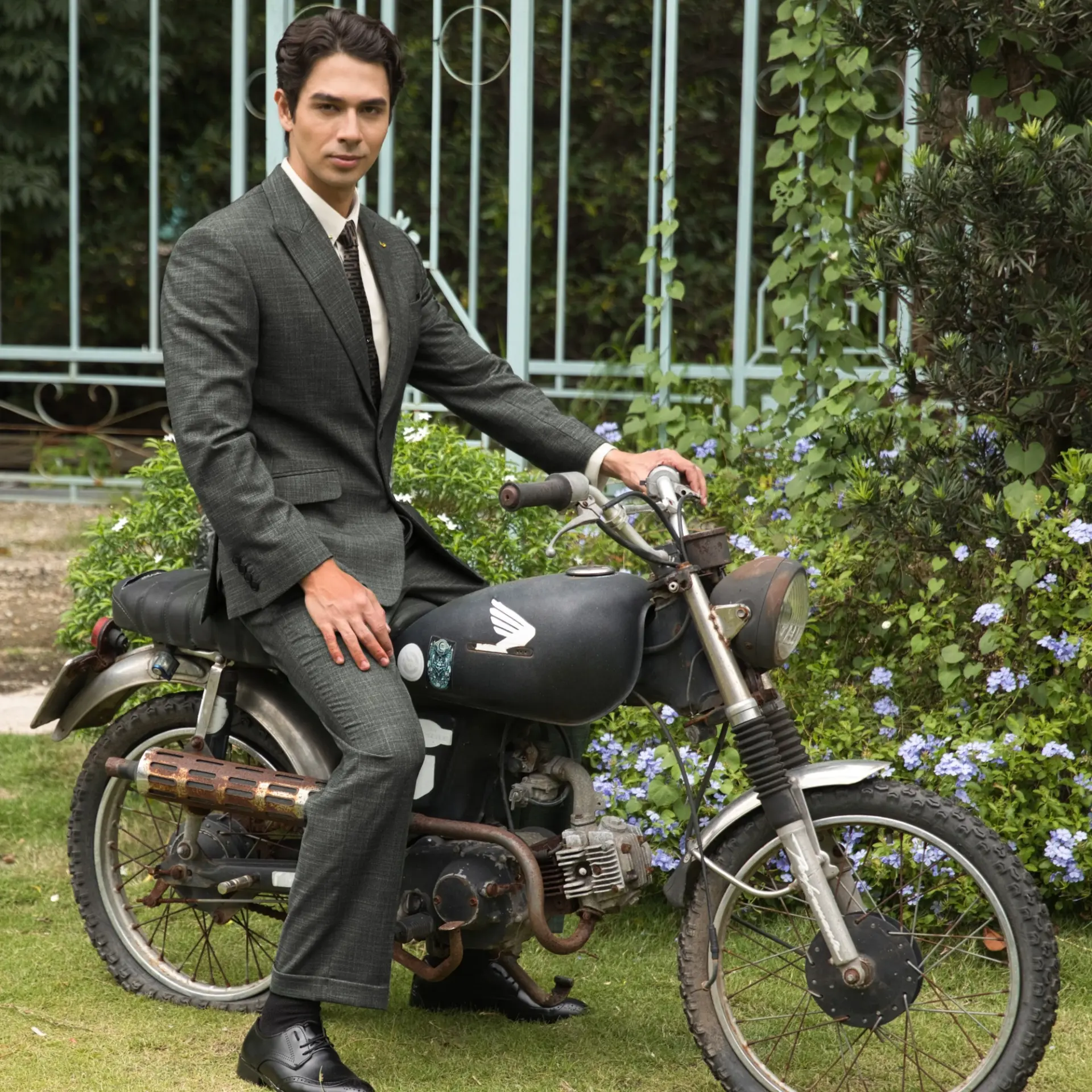 Man in a suit sitting on a vintage motorcycle in a garden setting with flowers in the background.