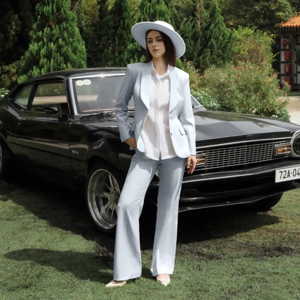 Woman in a stylish light blue suit and hat posing beside a vintage black car in a garden setting.
