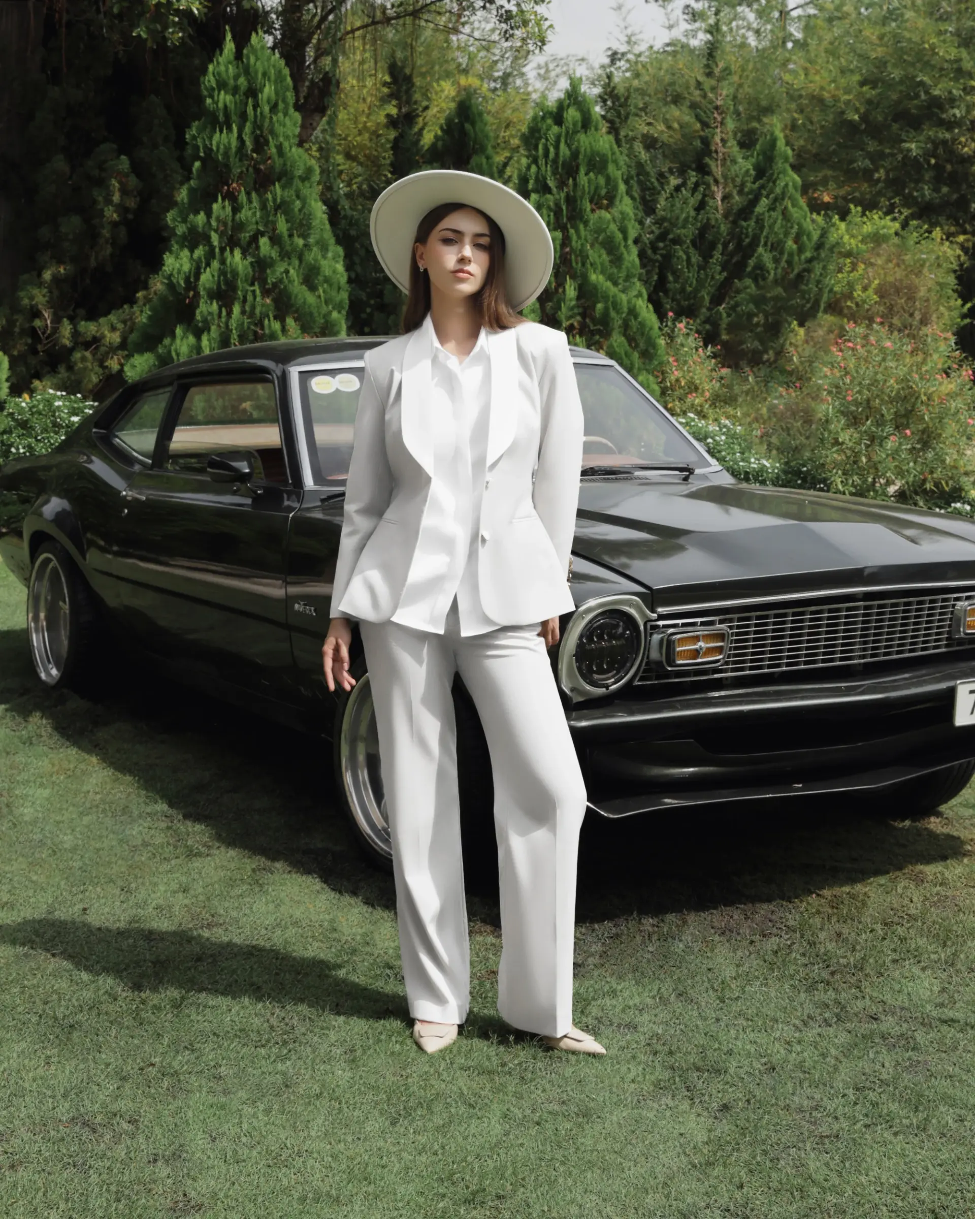 Person in a stylish white suit and hat stands by a classic black car in a lush garden setting.