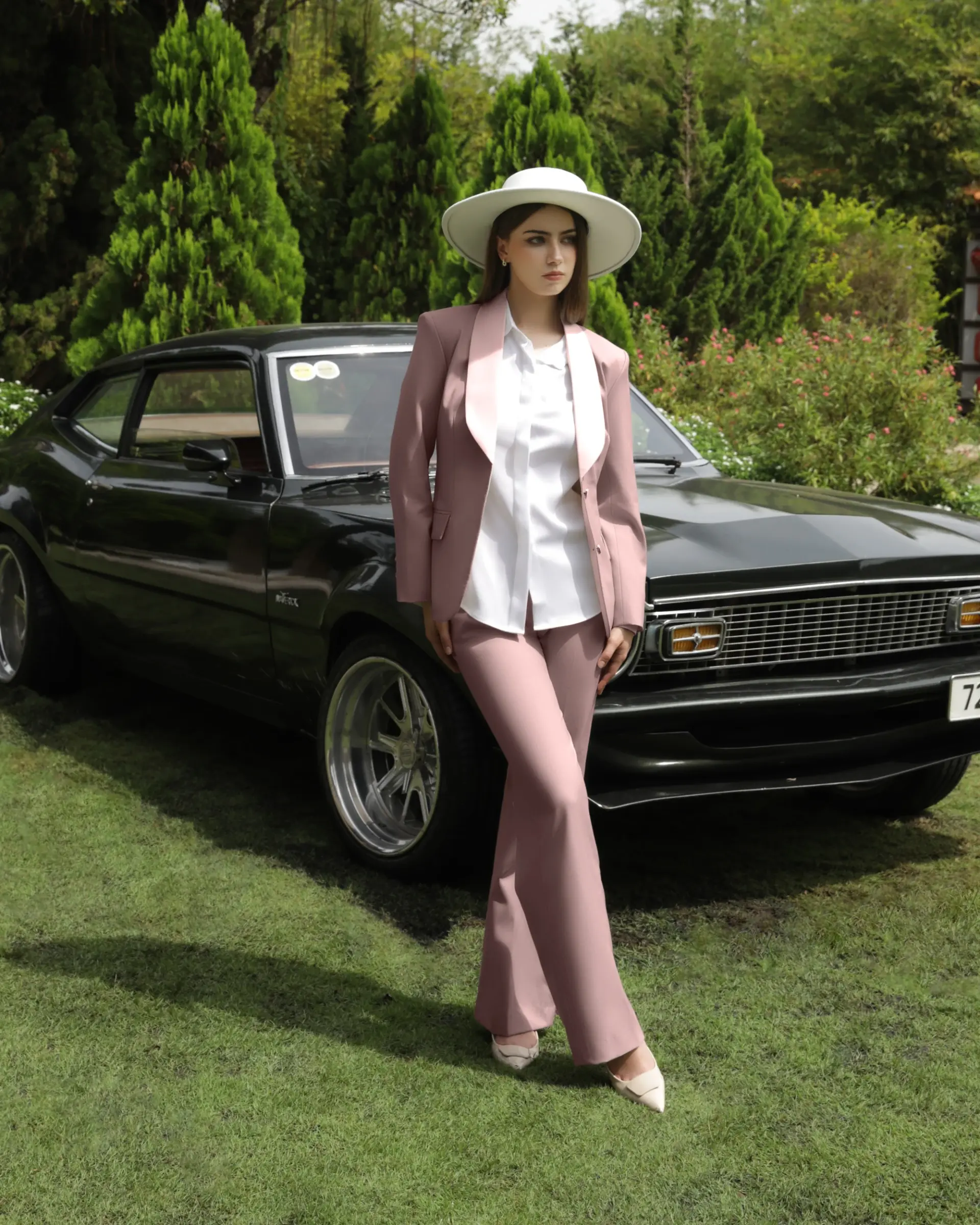 Woman in pink suit and wide-brimmed hat stands confidently beside vintage black car in a lush garden setting.