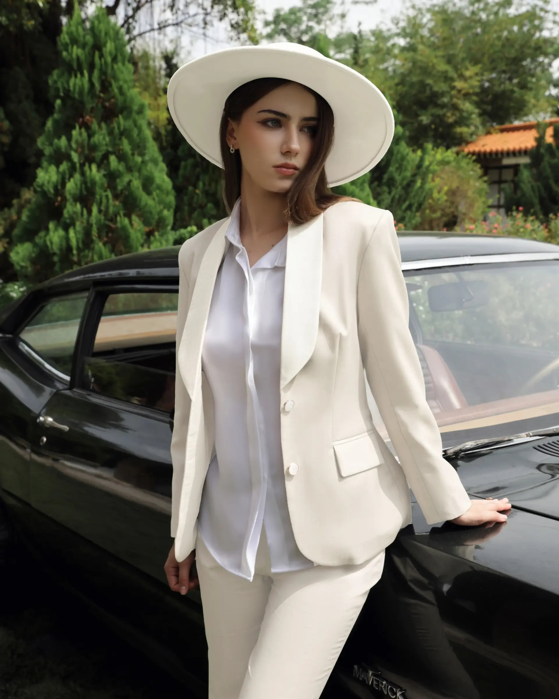 Woman in white suit and hat leans on black vintage car outdoors.