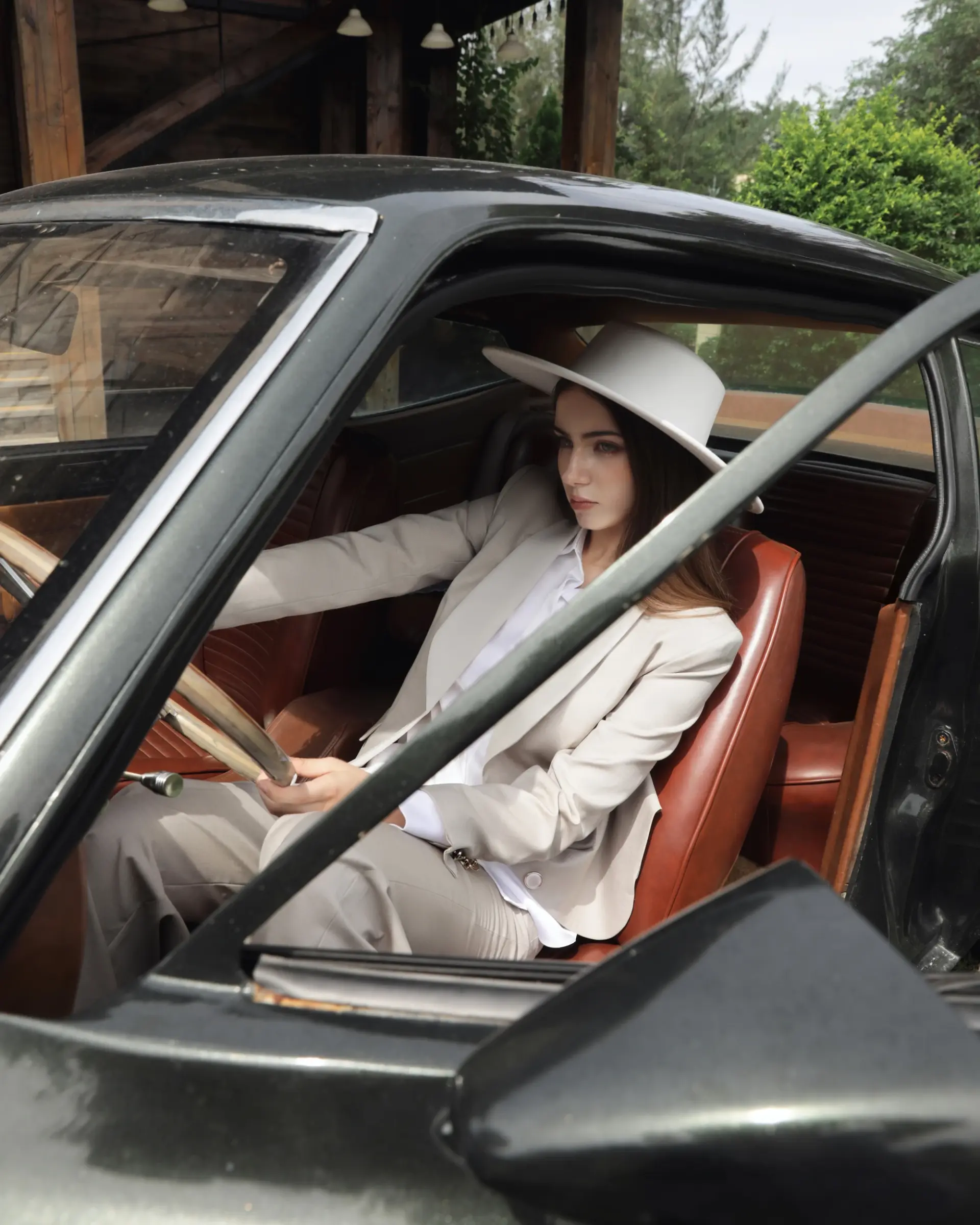 Woman in a stylish suit and hat driving a vintage car, showcasing fashion and classic automotive elegance.