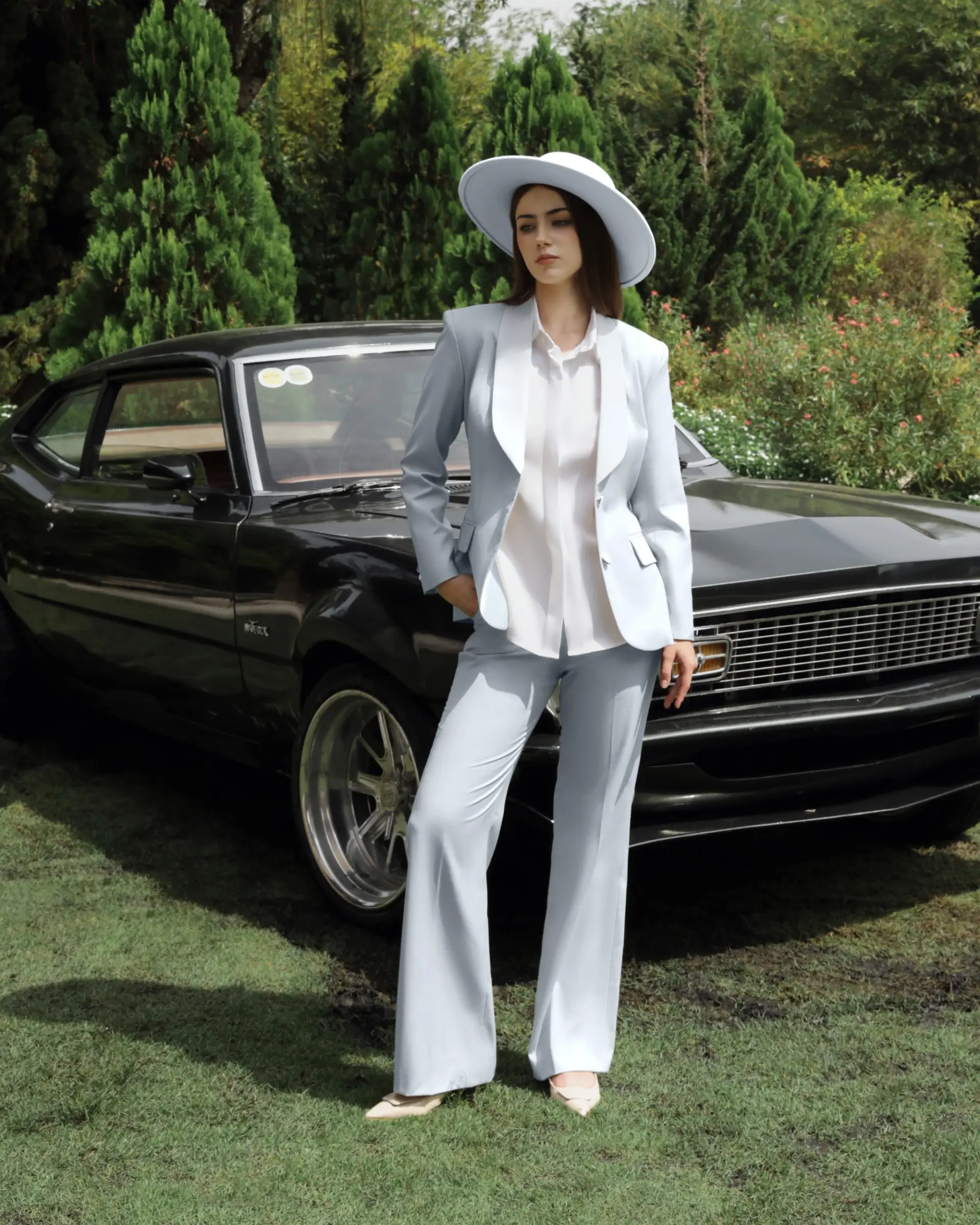 Woman in light blue suit and hat poses by vintage black car in a scenic garden setting.