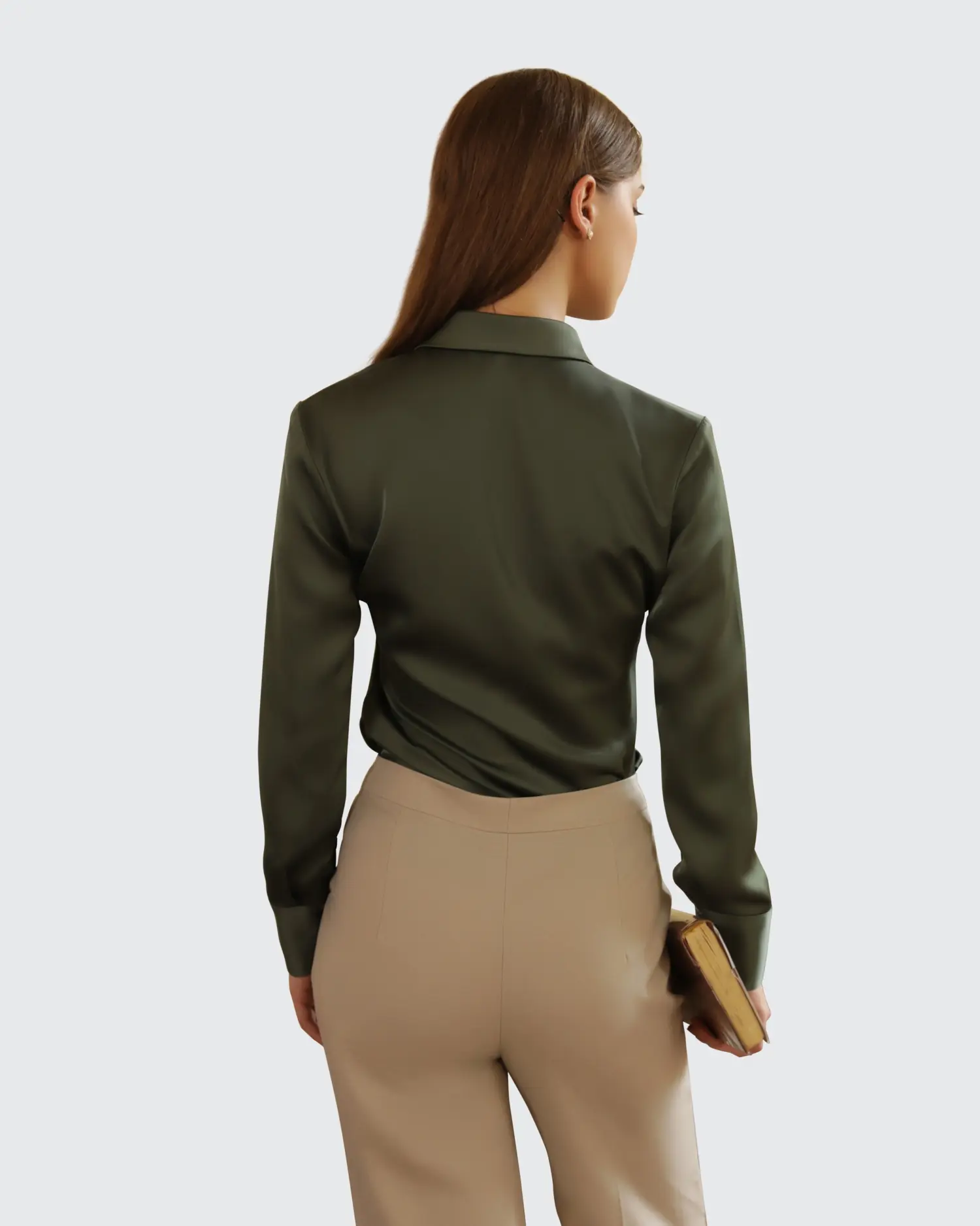 Woman in green blouse and beige pants holding a book, viewed from behind, on a plain background.