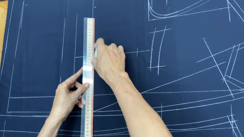 Hands using a ruler on fabric with white chalk marks, drafting a pattern for sewing.