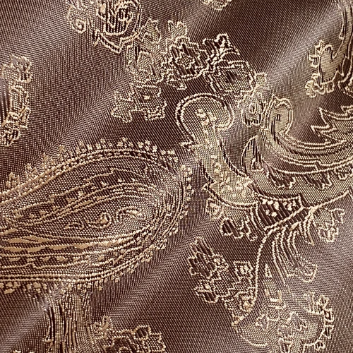 Close-up of intricate paisley fabric with gold embroidery on a brown background, showcasing elegant textile patterns.