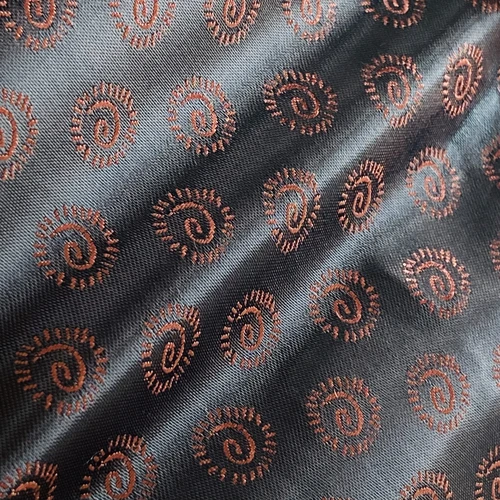 Close-up of fabric with an intricate spiral pattern, featuring black background and copper embroidery design.