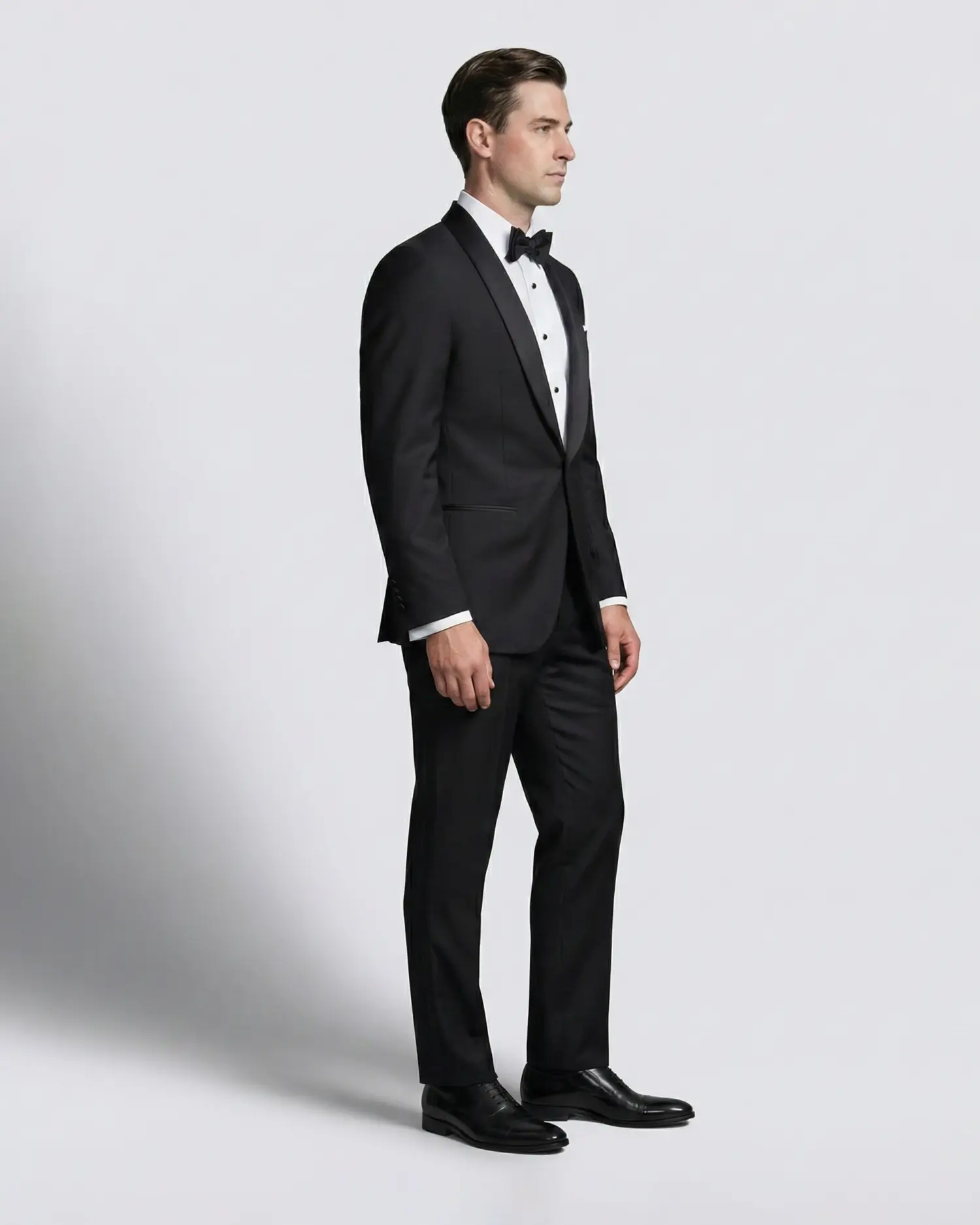 Man in a black tuxedo with bow tie, standing against a plain background for formal wear fashion.