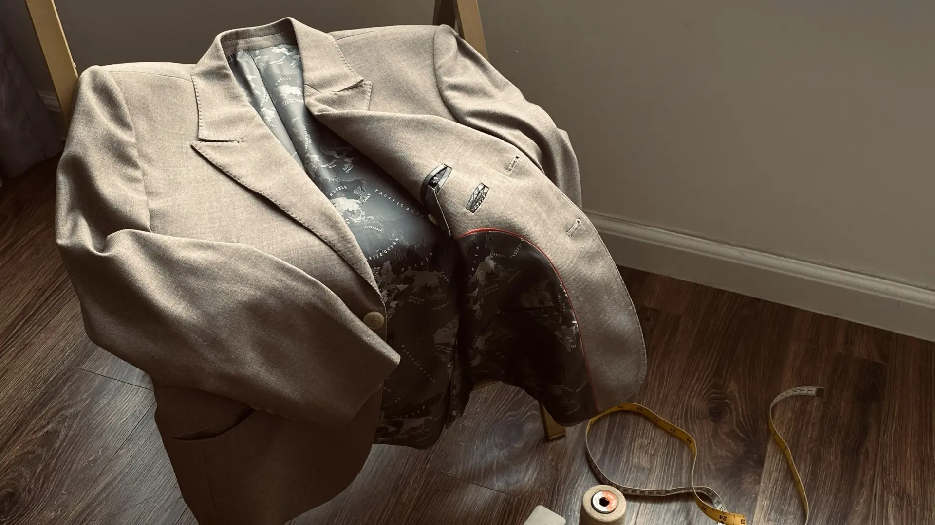 Brown blazer on chair, detailed interior lining visible, with measuring tape and thread spool on hardwood floor.