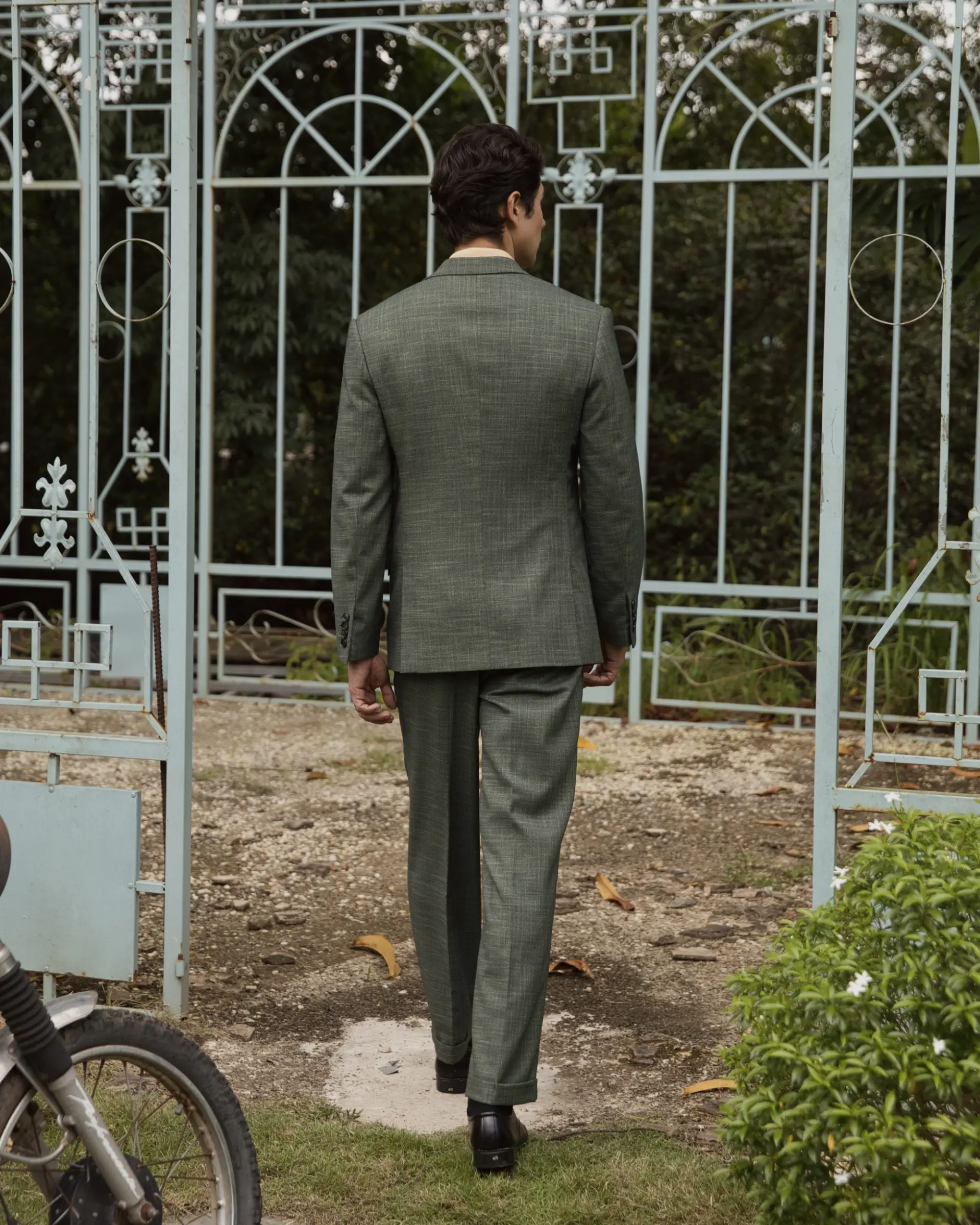 Man in green suit walking through garden gate, motorcycle in foreground.
