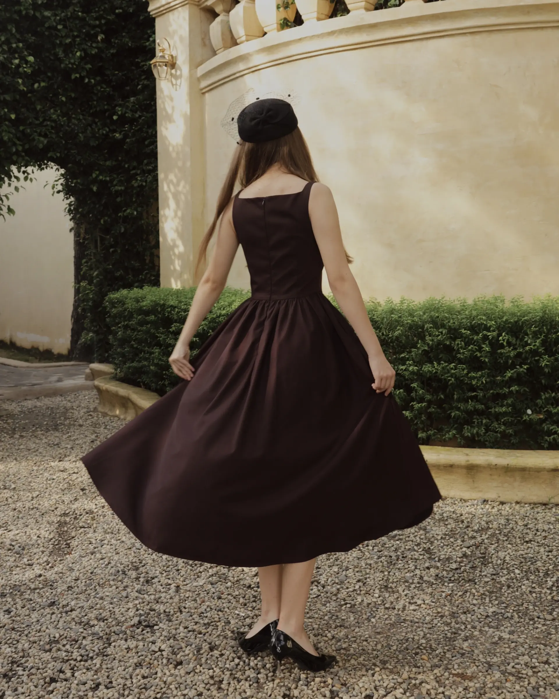 Woman in brown dress twirls on pebbled path near garden wall, wearing black beret. Vintage style fashion photography.