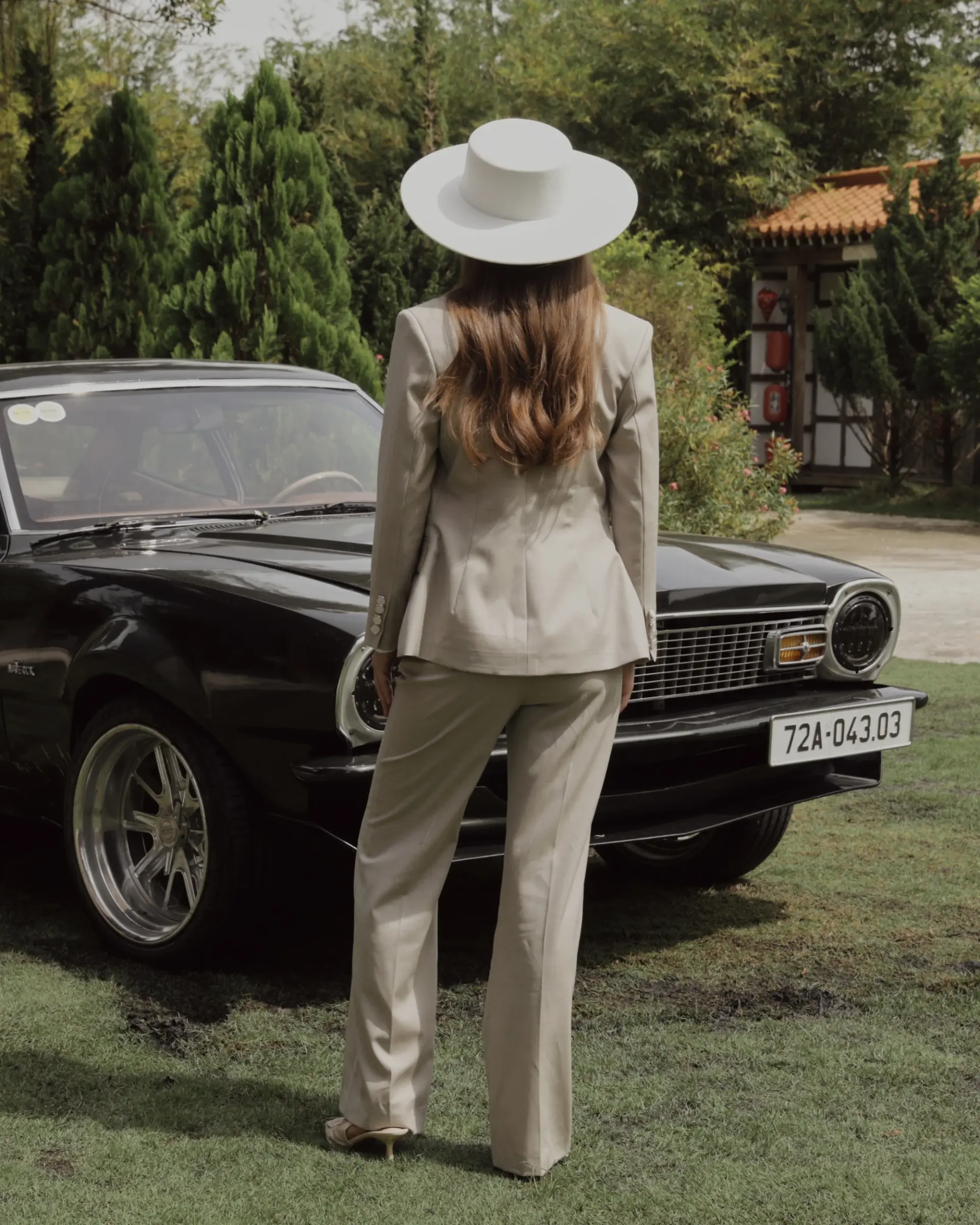 Woman in beige suit and white hat standing by a classic black car on a grassy driveway.