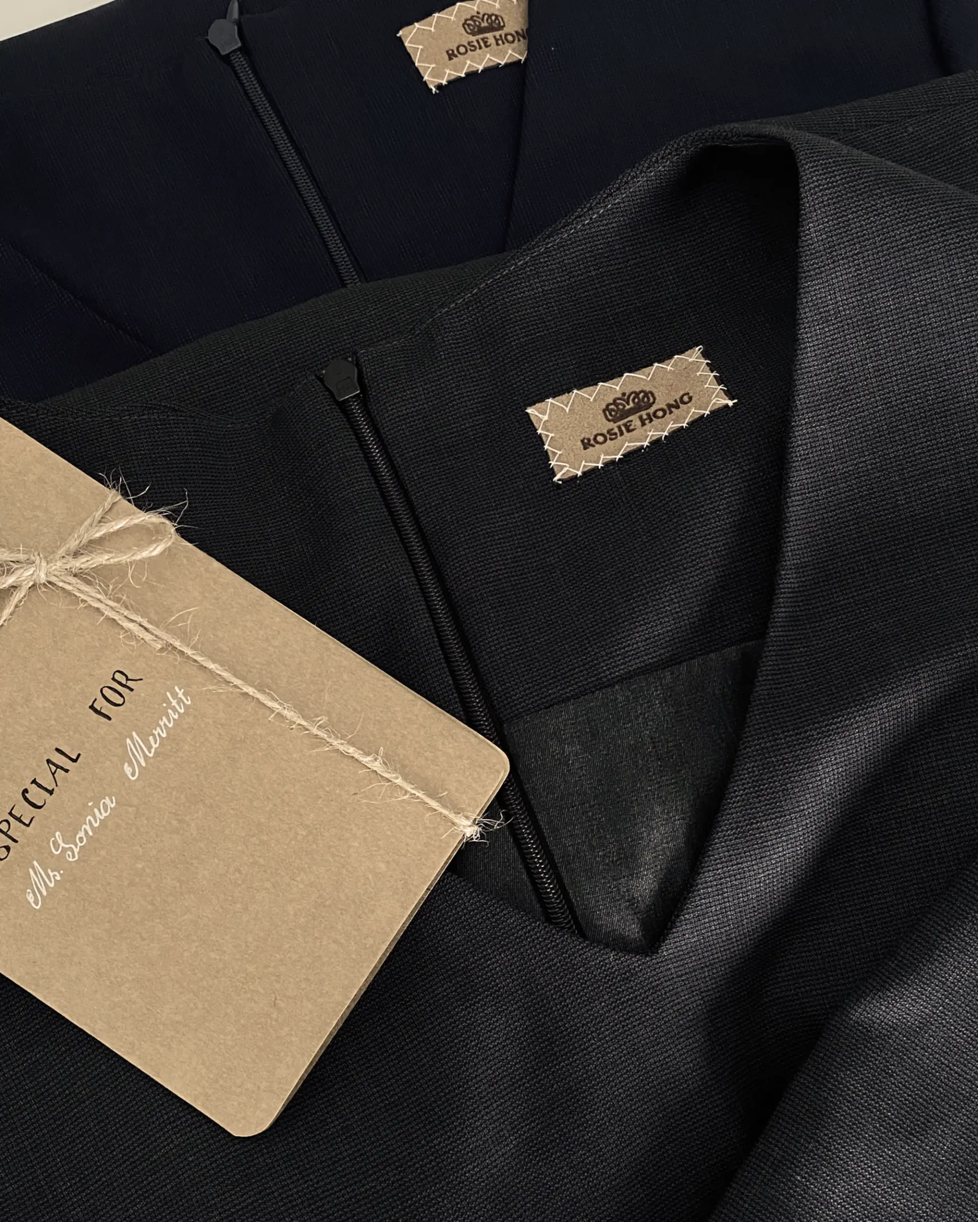 Close-up of two dark garments with Rosie Hong labels and a personalized tag attached with string.