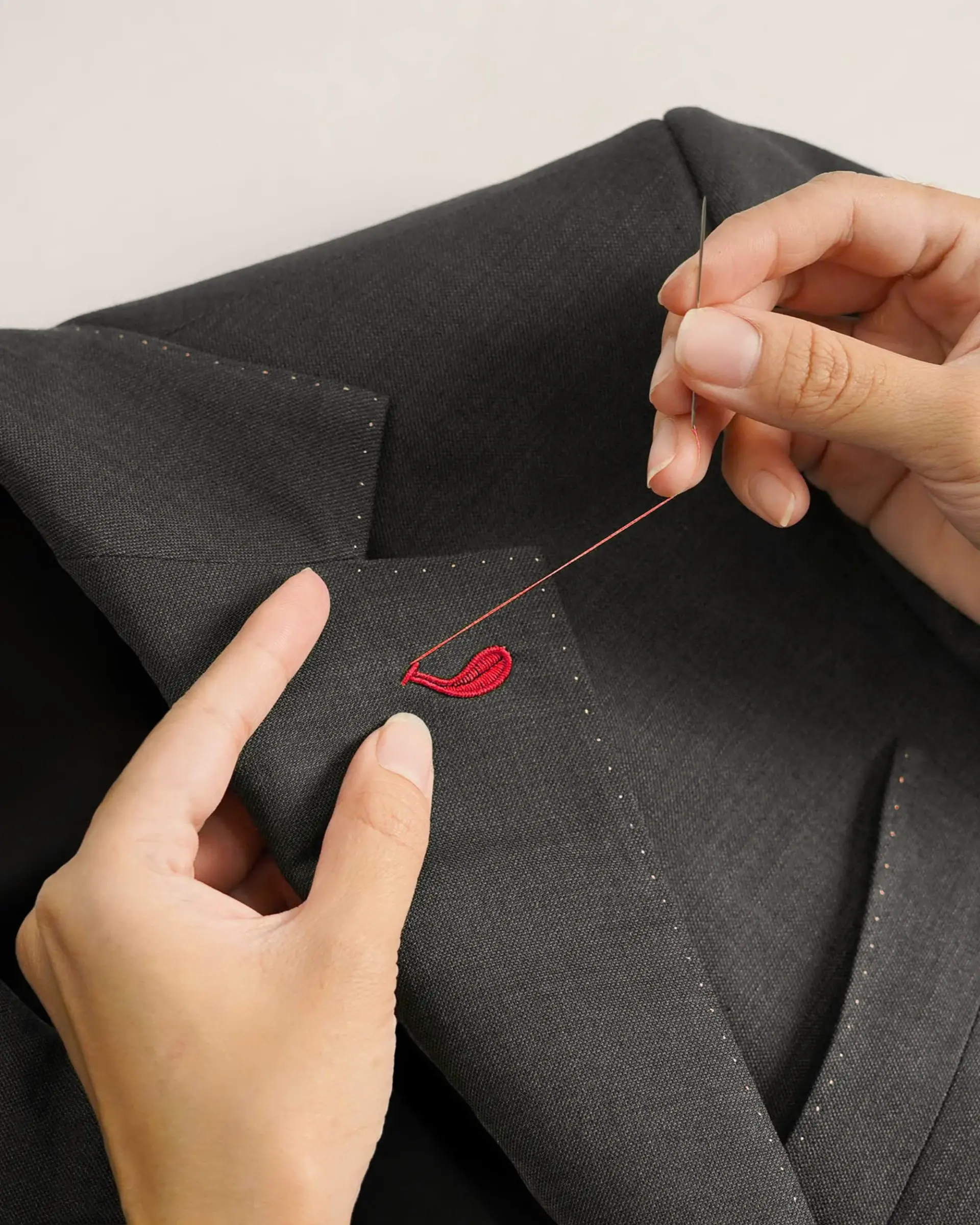 Person hand-stitching red buttonhole on a dark suit jacket using a needle and thread.