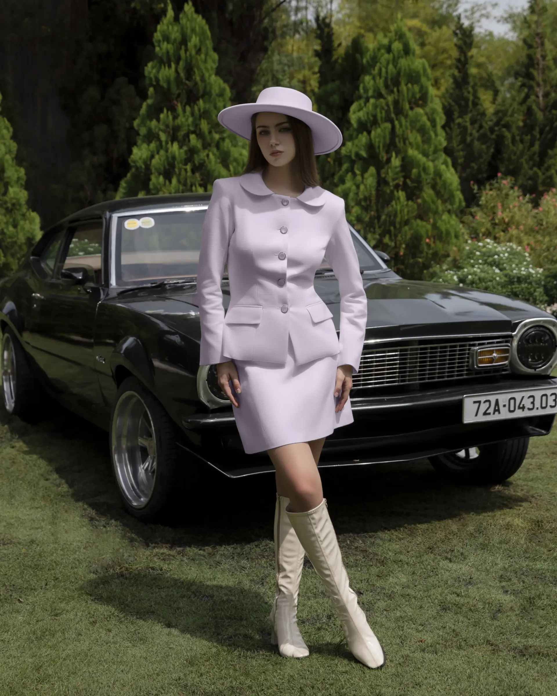 Fashionable woman in lavender outfit and hat posing by classic car in a lush garden setting.