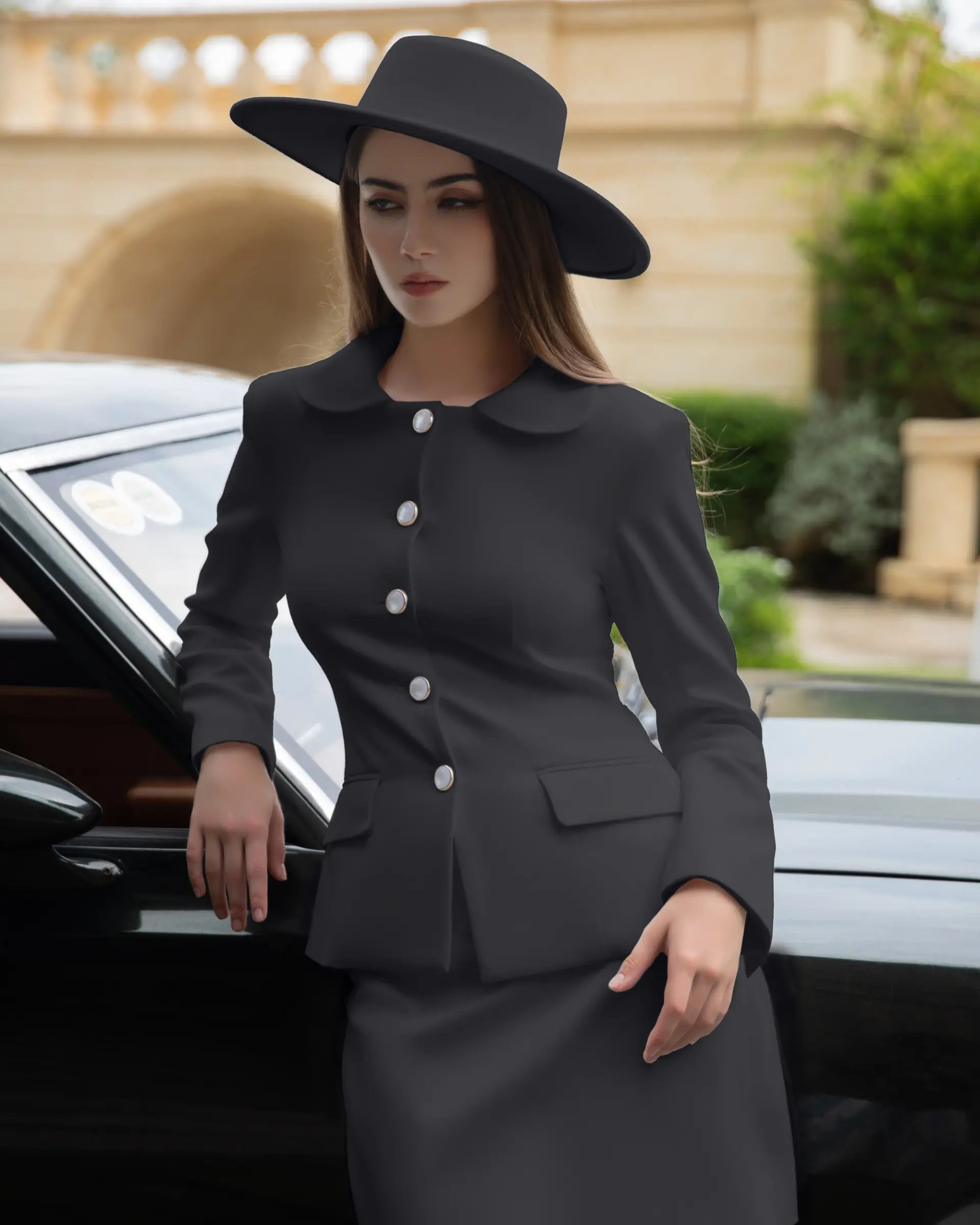 Woman in elegant black suit and wide-brimmed hat leaning on car, outdoor setting.