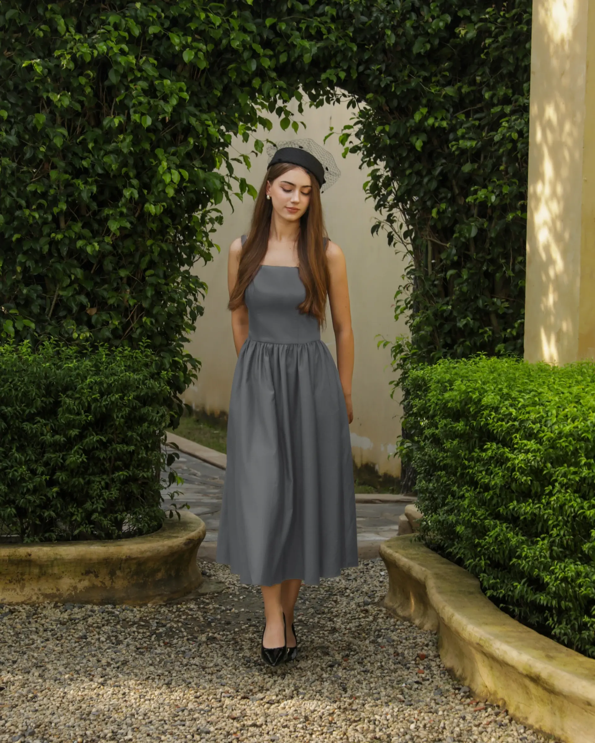 Woman in a gray dress and black hat walking in lush garden path.