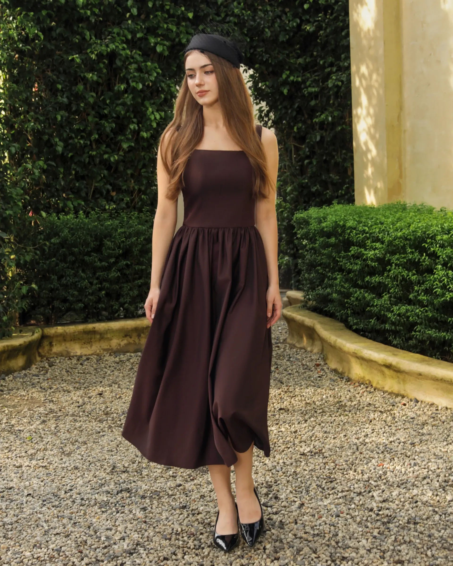 Woman in elegant brown dress and black hat walking on gravel path in garden.