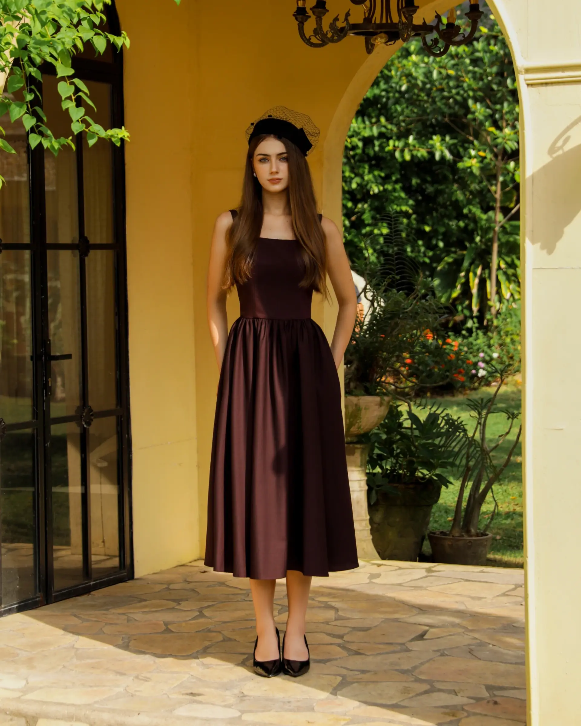 Elegant woman in a dark dress and hat standing in a garden archway, exuding classic style and charm.