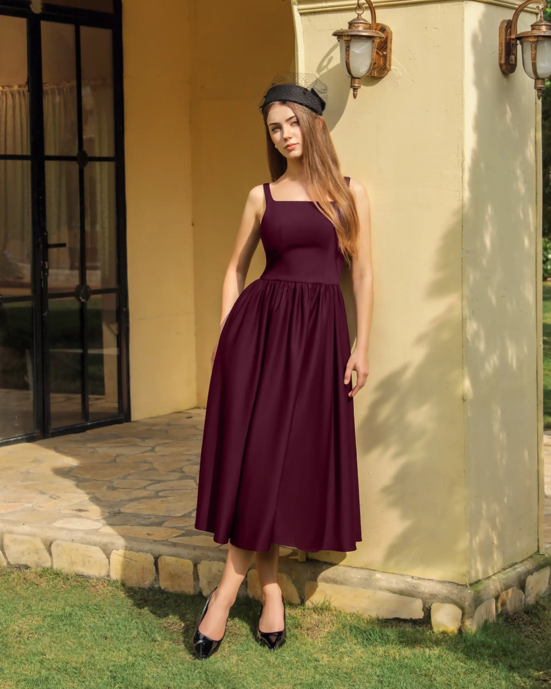 Woman in a burgundy dress and black hat stands by a sunny yellow wall and a glass door, exuding elegance and style.