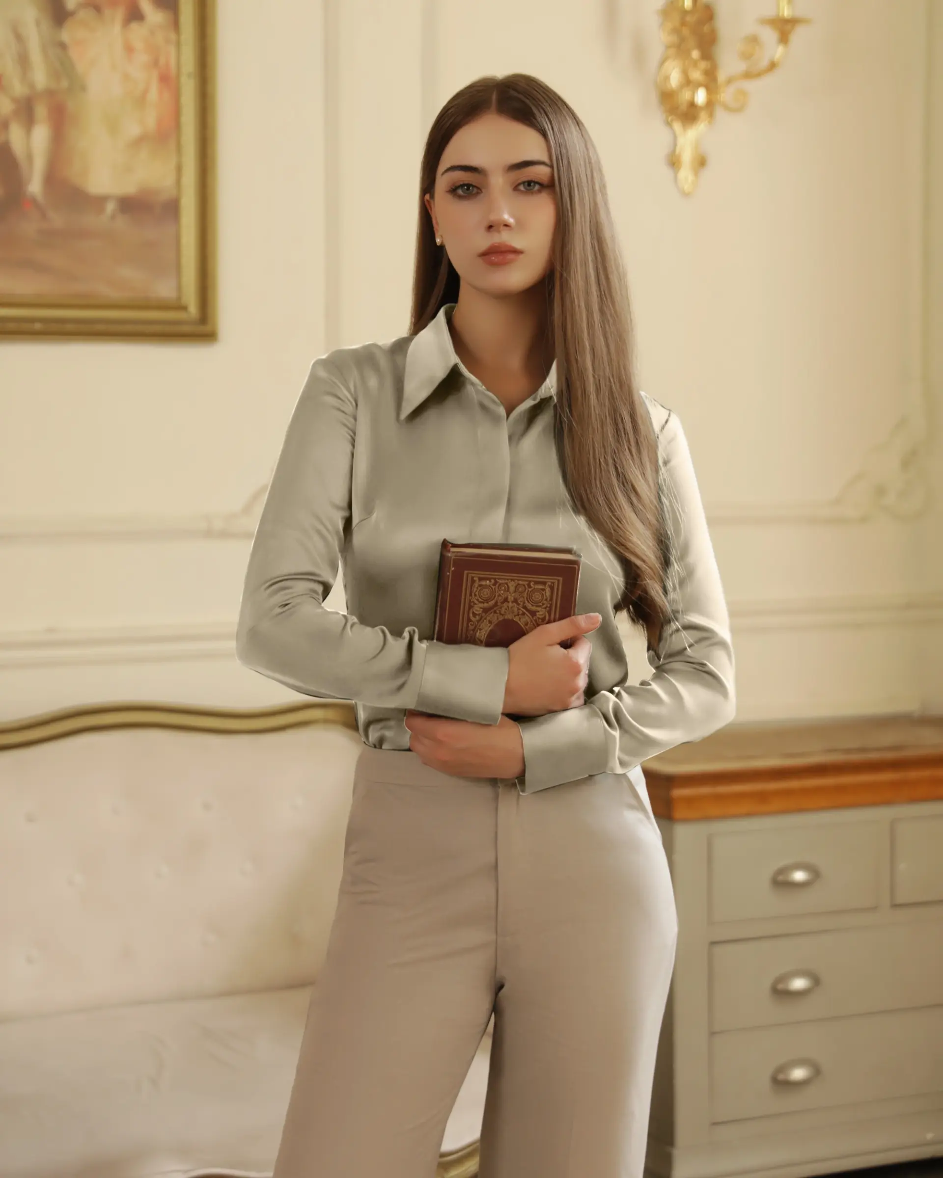 Woman holding an ornate book in an elegant room, wearing a satin blouse and light pants.
