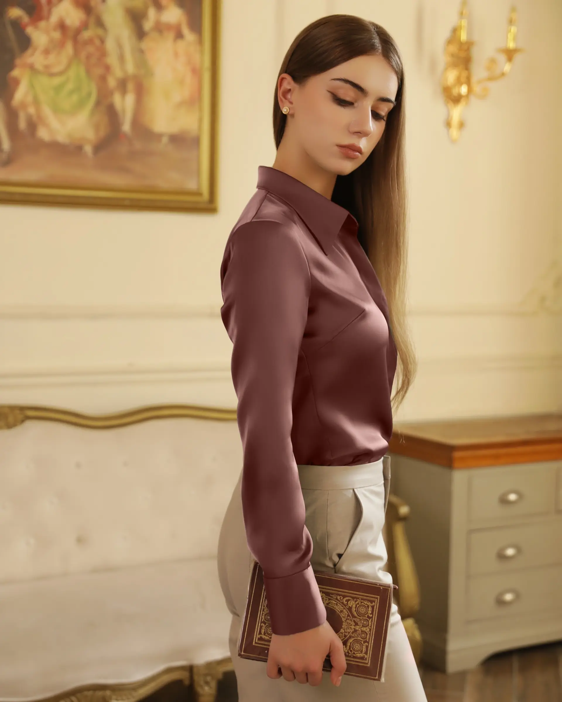 Elegant woman in satin blouse holding a book, standing in a vintage room with classic decor.